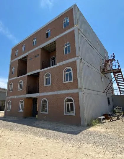A four-story building under construction with brick and shipping container sections, outdoor metal staircase on the right side, and clear blue sky.
