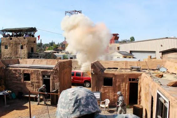 Military personnel throwing a grenade or explosive device in a desert-like environment with buildings, smoke, and a red vehicle.