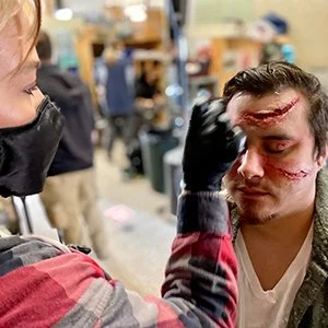A makeup artist applies special effects makeup with scars to a man's face in a workshop setting.