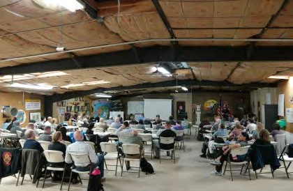 People attending a presentation or meeting in a large, open room with a projector screen at the front and a mural on the wall.