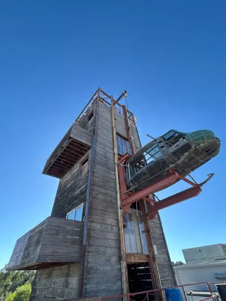 A tall wooden tower with a vintage aircraft attached to its side, set against a clear blue sky.