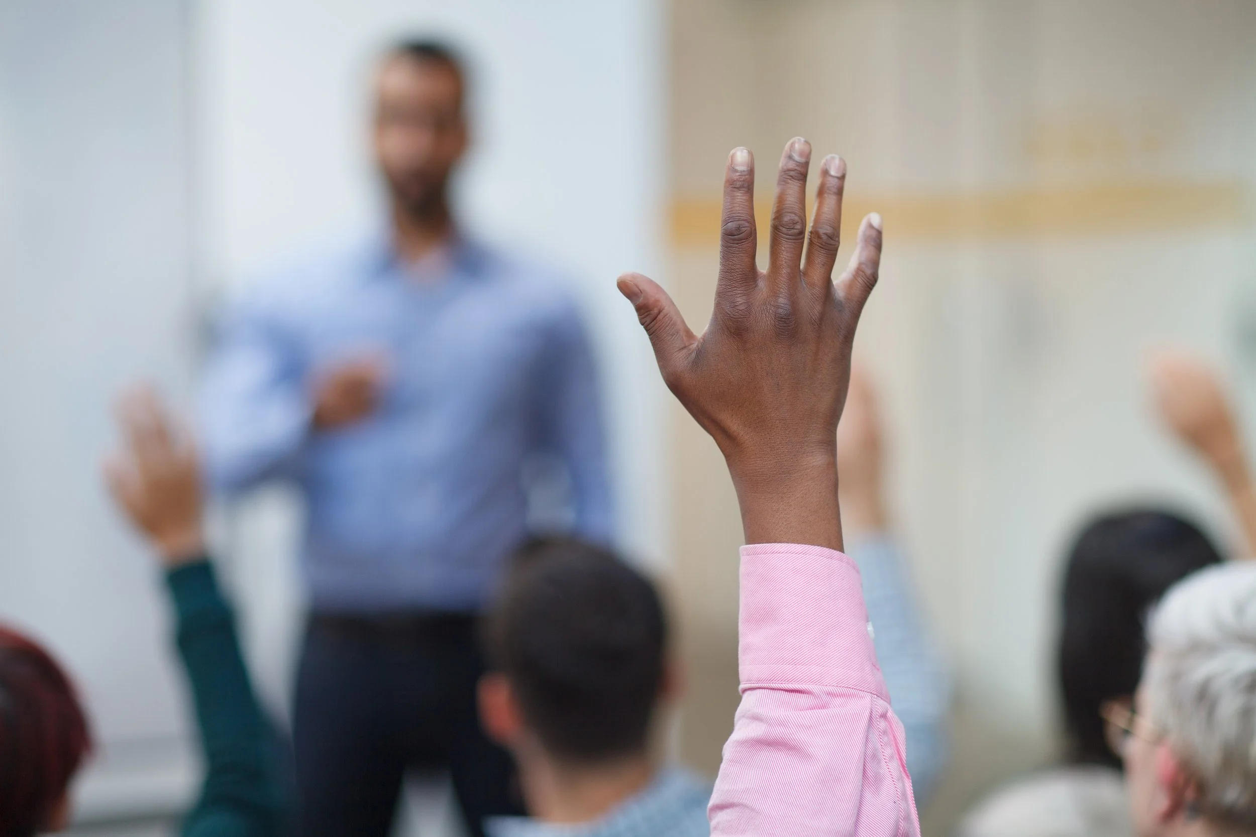 Child raising hand in classroom, teacher in background, students seated, classroom setting