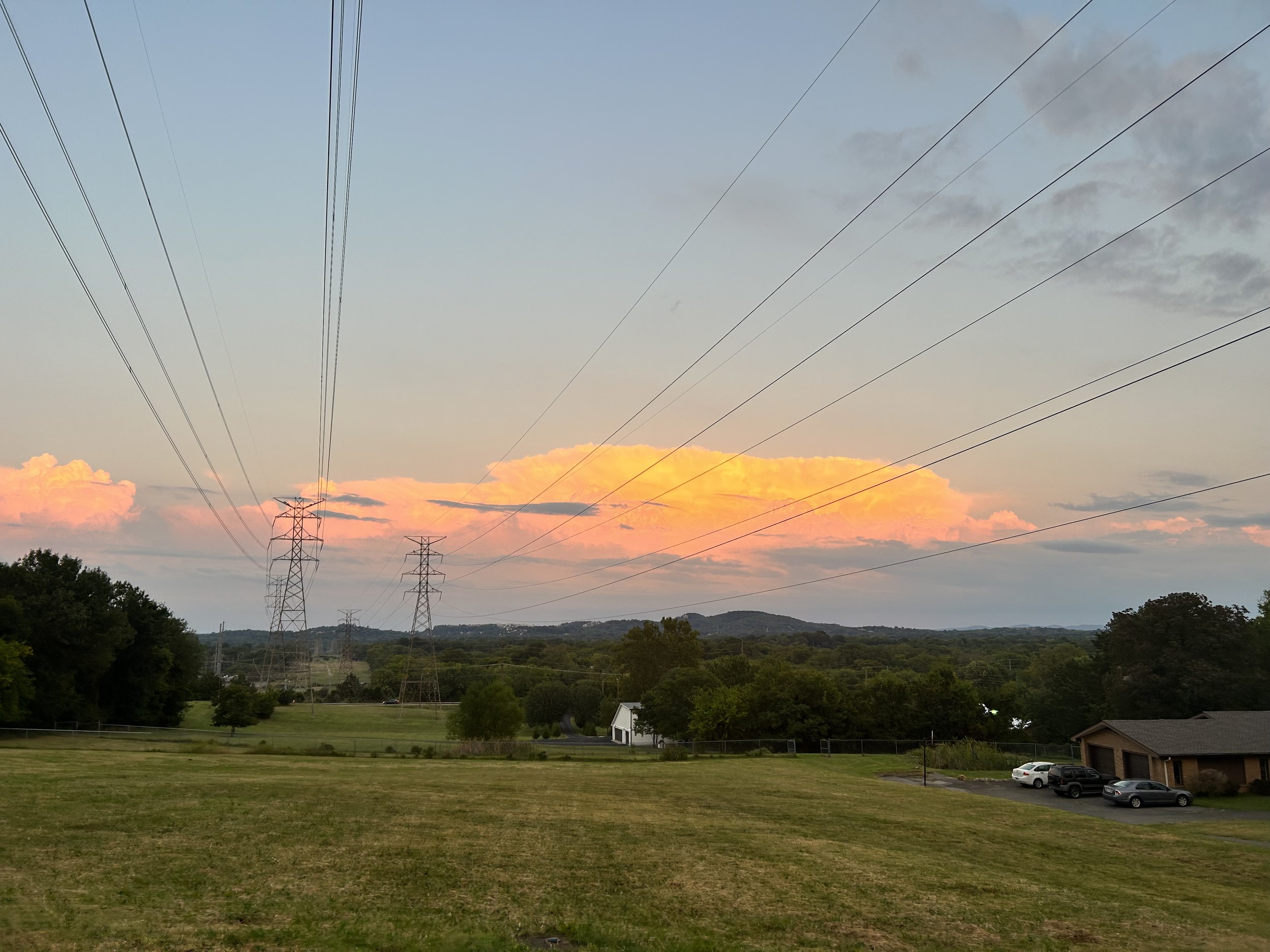 Landscape scene with multiple power line towers and lines stretching across the sky, along with a grassy field, trees, houses, and a parking lot with cars. The sky is partly cloudy with pink and orange hues from the setting sun.
