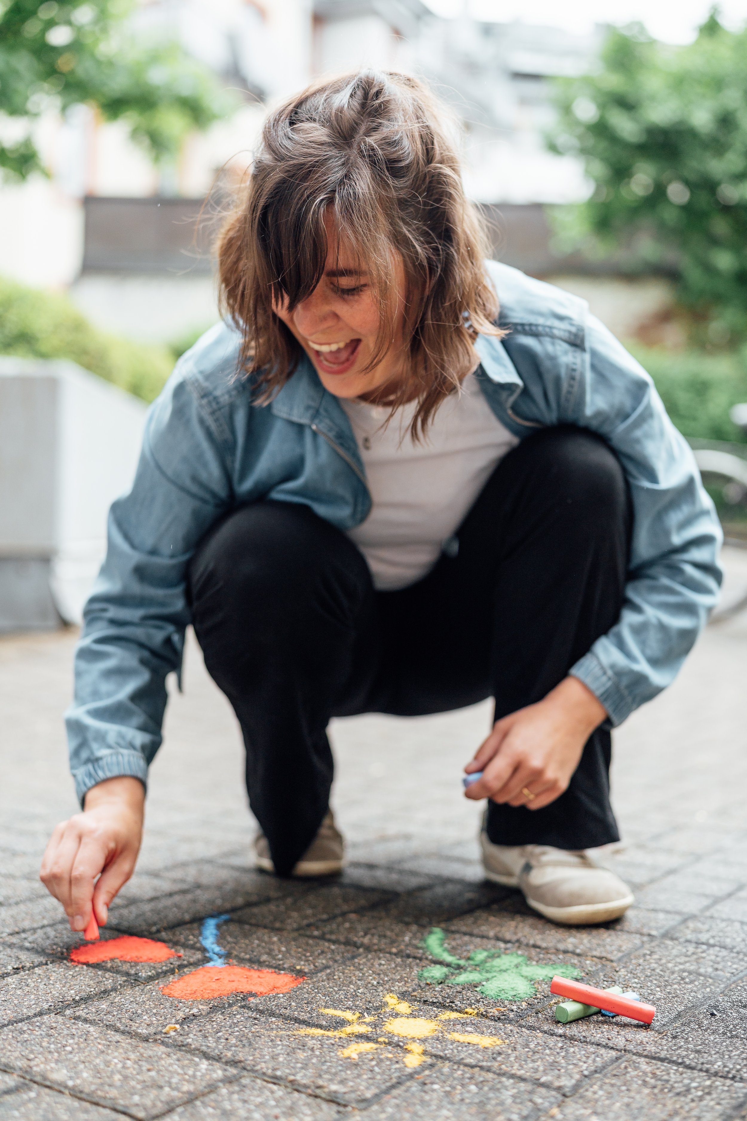 Eine junge Frau, die auf einer gepflasterten Fläche bunte Kreidezeichnungen auf die Erde zeichnet. Sie lächelt und scheint Spaß daran zu haben, mit den Kreidefarben zu spielen.