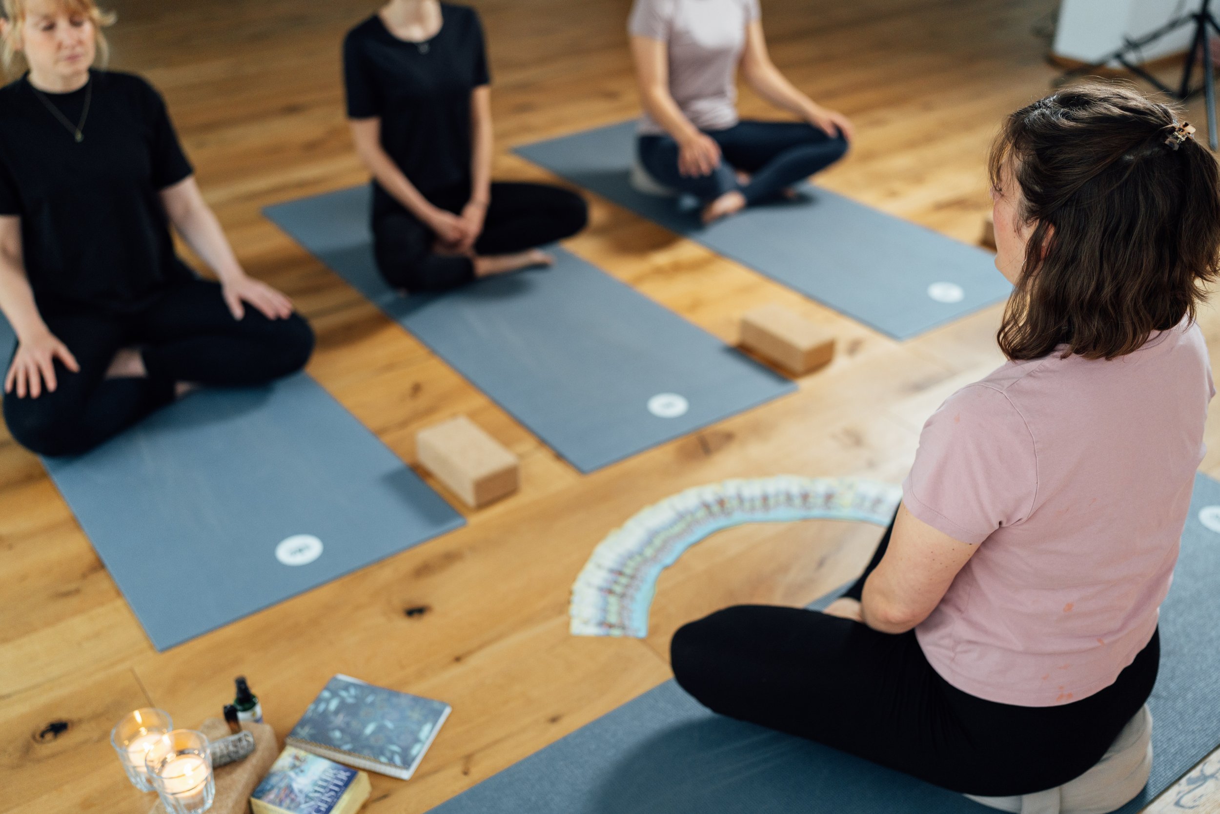 Vier Frauen sitzen im Schneidersitz auf Yogamatten im Kreis in einem Raum mit Holzboden. Eine Frau hat braune, lockige Haare und trägt eine rosa Bluse. Die anderen Frauen tragen dunkle Kleidung. Auf dem Boden vor ihnen liegen Bücher, eine Kerze, ein Schraubglas mit Pipette, ein Notizbuch und verschiedene Karten oder Papierstreifen in Regenbogenfarben.