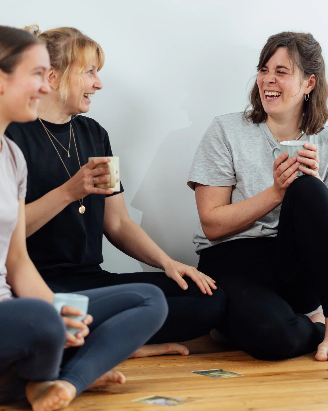 Gruppe von Frauen sitzt im Kreis auf dem Boden und trinkt Kaffee, sie lachen und unterhalten sich in einem hellen Raum.