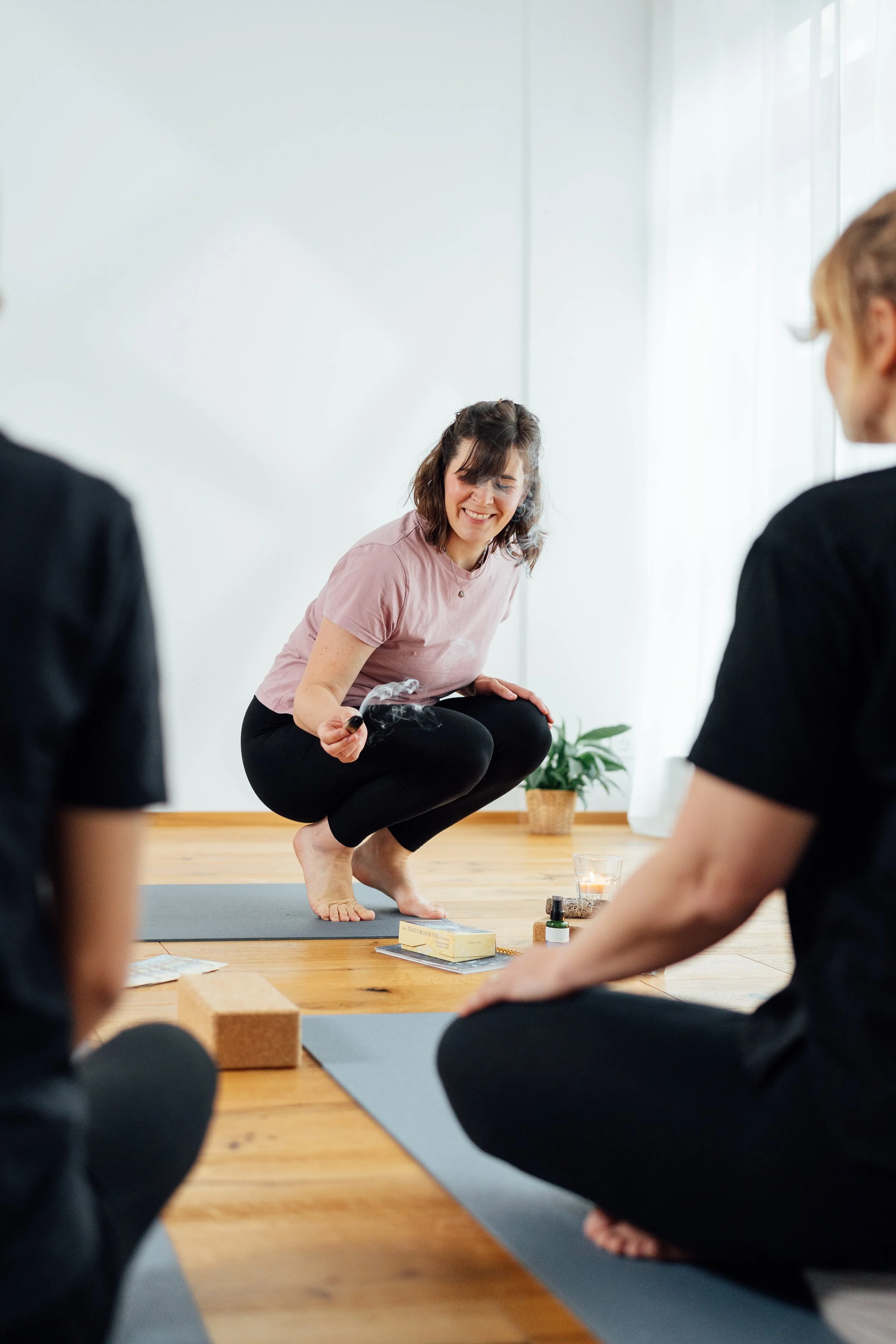 Eine Frau im Yogakurs, die in der Hocke auf einem Yoga- oder Pilates-Matte sitzt, während sie eine Trainingseinheit mit mehreren Teilnehmern im Hintergrund durchführt. Sie hat braune, gewellte Haare und trägt ein rosa T-Shirt. Es befinden sich verschiedene Yoga-Accessoires und Pflanzen im Raum.