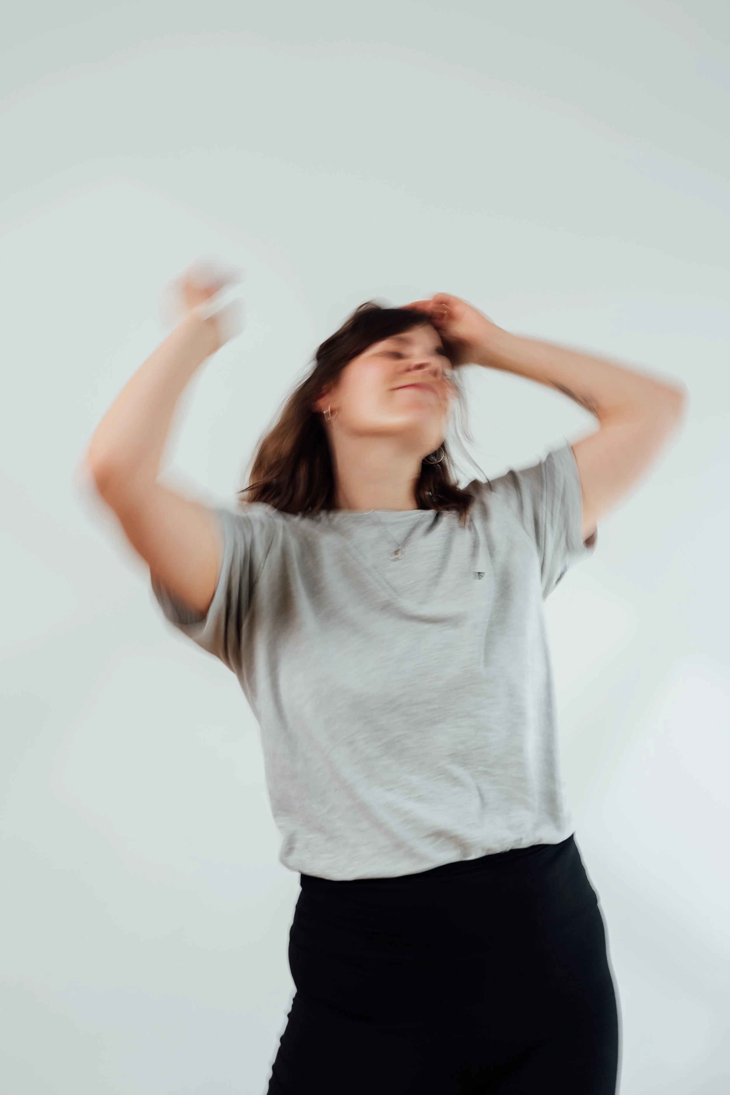 Eine junge Frau mit braunen Haaren, die in einem grauen T-Shirt und schwarzen Hosen tanzt, während sie die Augen schließt und die linke Hand an den Kopf legt. Sie steht vor einem weißen Hintergrund.