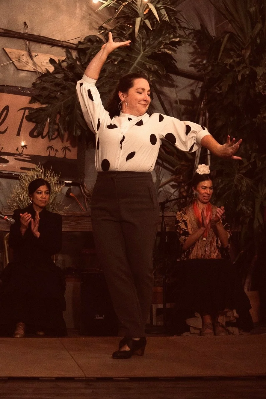 A woman dancing on stage with her eyes closed, wearing a white blouse with black polka dots and black pants, while two women sit behind her clapping, in a warm, tropical-themed setting.