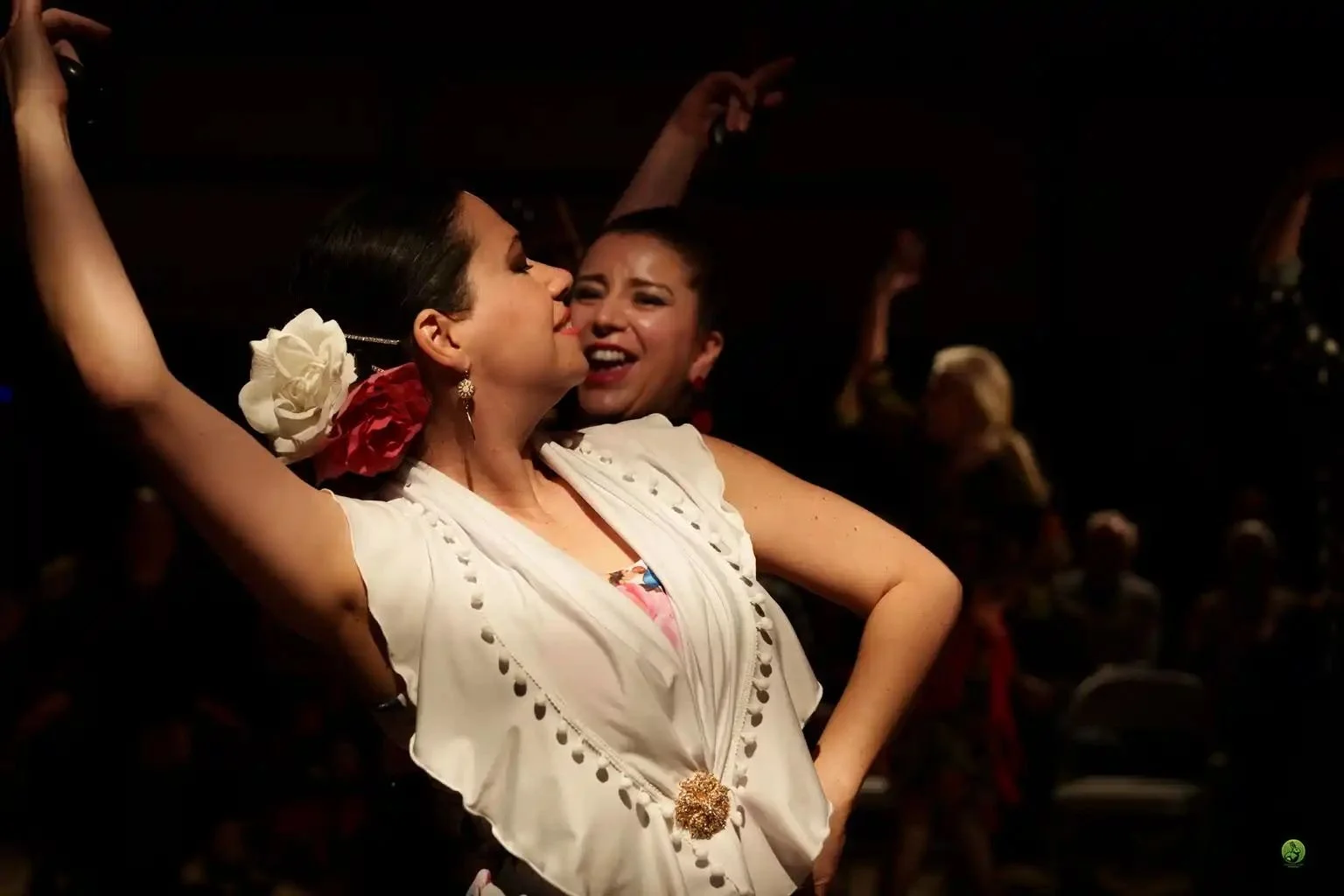 Two women dancing closely together at a flamenco show, one woman with dark hair wearing a traditional white embroidered dress with a large flower in her hair, and the other woman smiling behind her.