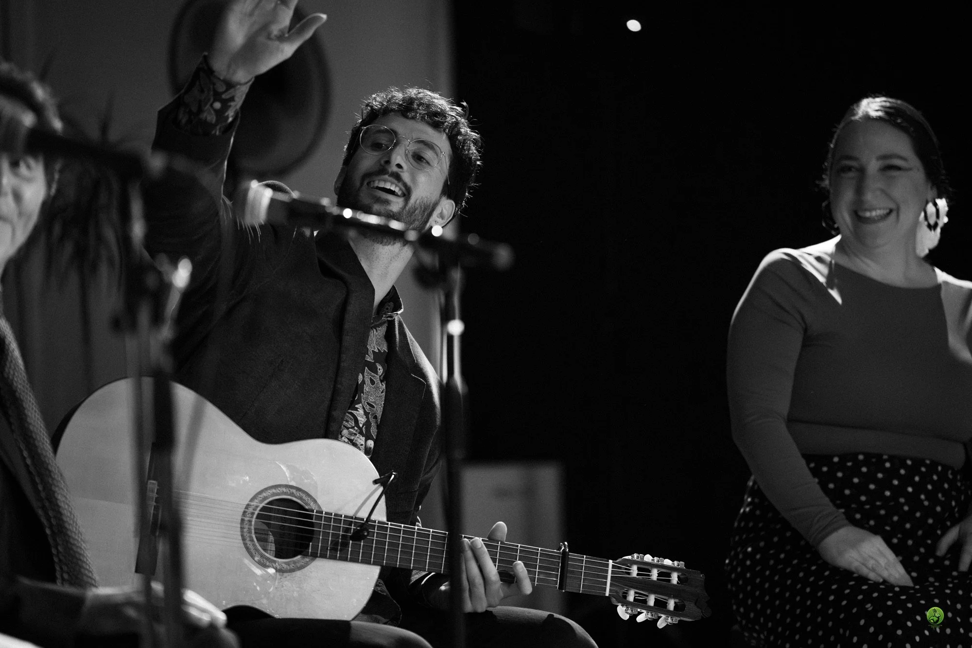 Black and white photo of a man with curly hair and glasses playing an acoustic guitar, smiling, while a woman with short dark hair and earrings looks on, all seated, during a flamenco performance.