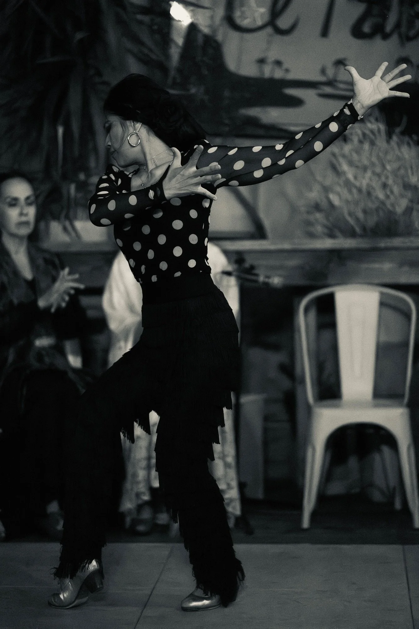 A woman dancing indoors wearing a black dress with white polka dots, fringed black pants, and metallic shoes. She has dark hair, large earrings, and is striking a dance pose with one arm extended and the other hand near her chest. In the background, 