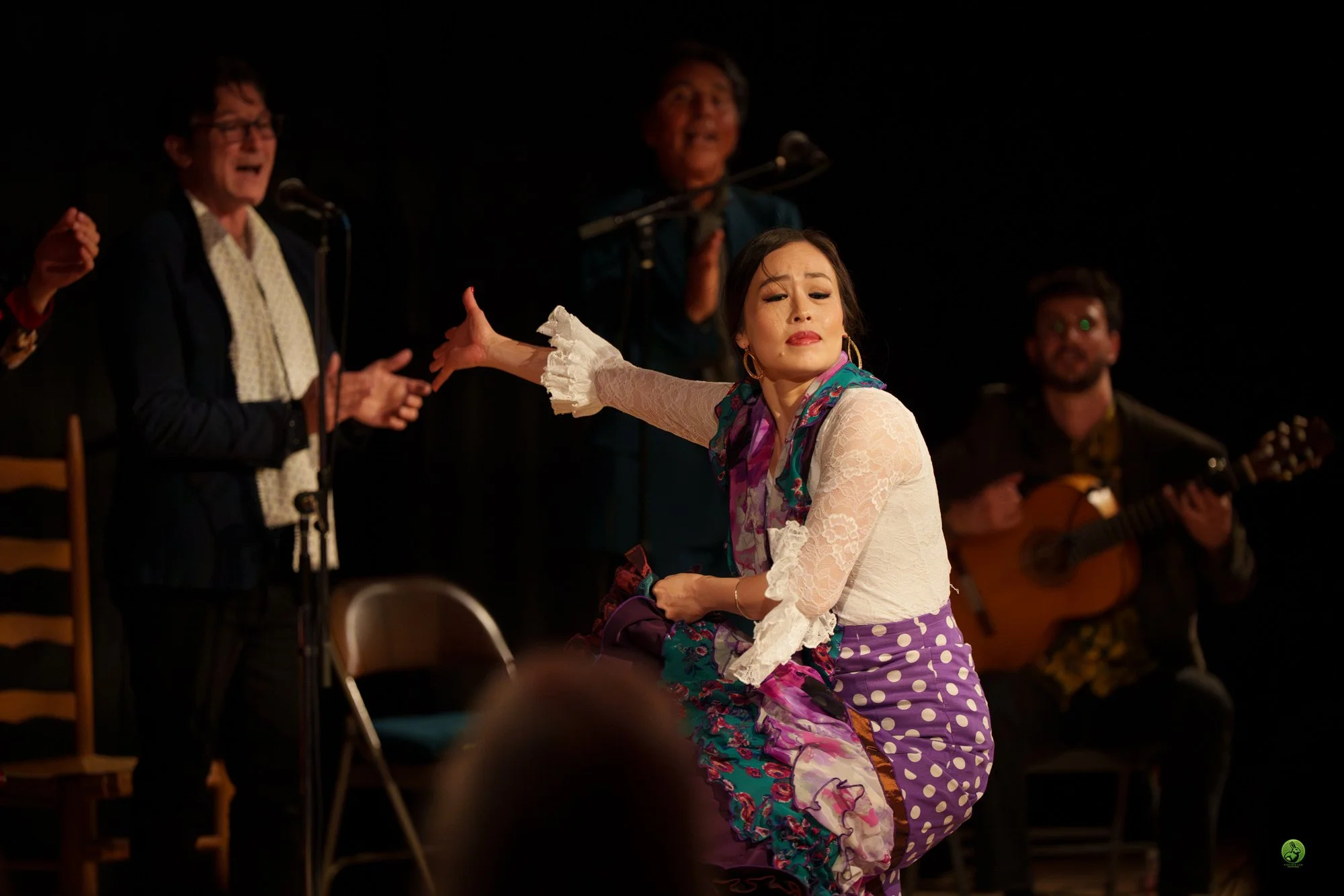 A woman in colorful flamenco clothing performs a dance on stage, with background singers and a guitarist. The woman has her eyes closed and one arm extended, showcasing expressive dance movements.