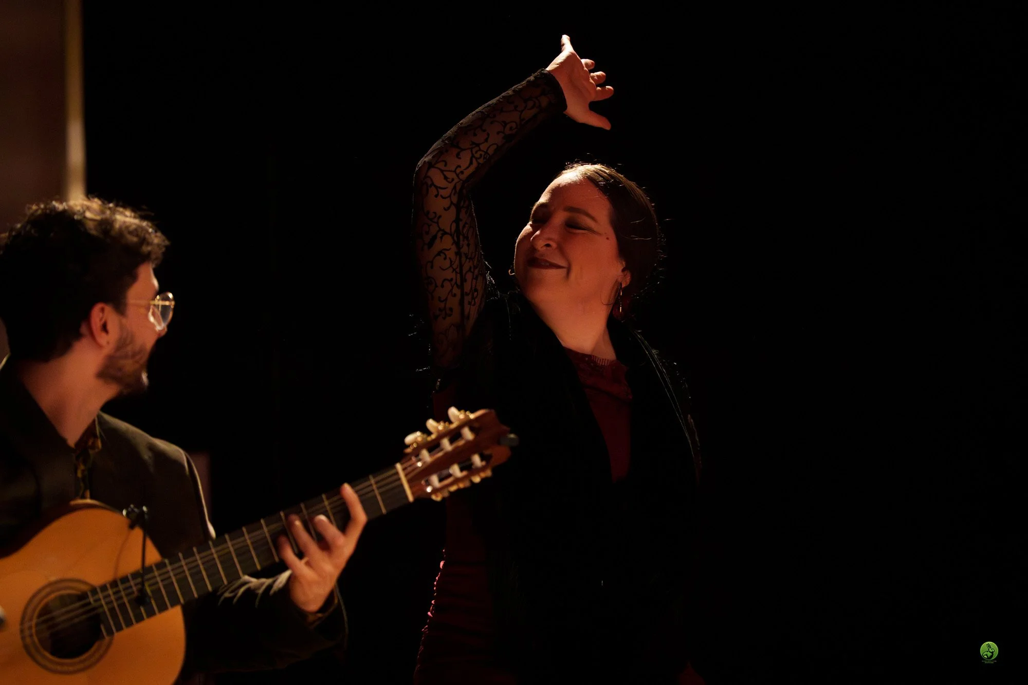 A woman with dark hair and earrings, wearing a black lace long-sleeve top, is dancing with her eyes closed and a joyful expression. Partially visible, a man with glasses and a beard is playing an acoustic guitar, wearing a dark shirt. The background 