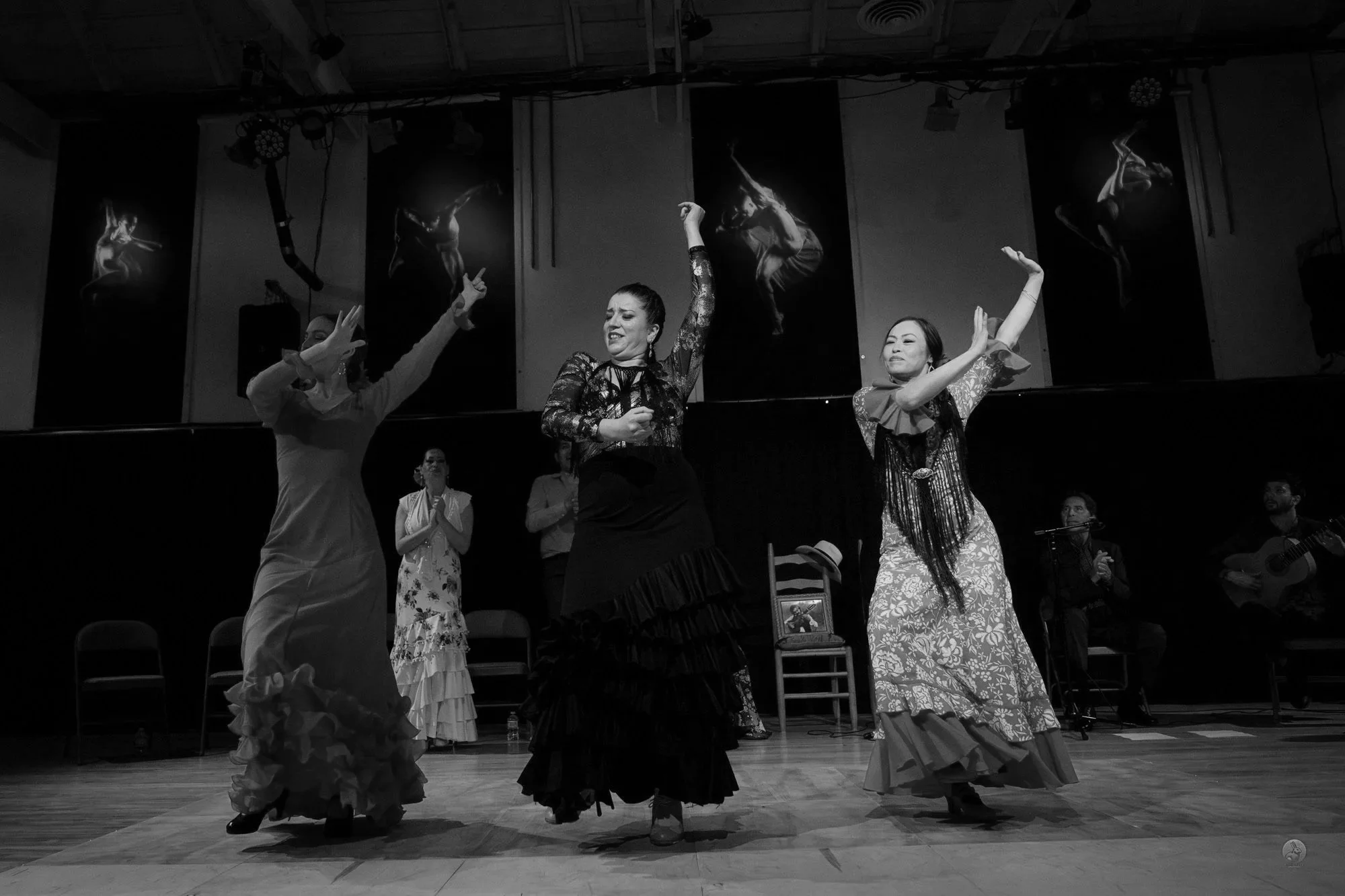 Women performing flamenco dance on stage with performers watching in the background. The women are dressed in traditional dresses with ruffles and are moving gracefully.