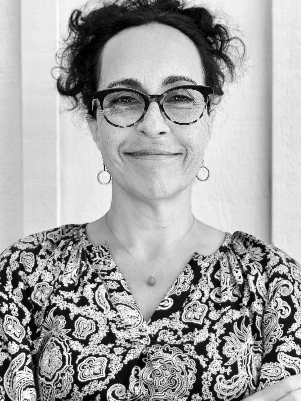 A black-and-white photo of a woman with curly short hair, wearing glasses, earrings, and a paisley patterned top, smiling at the camera.