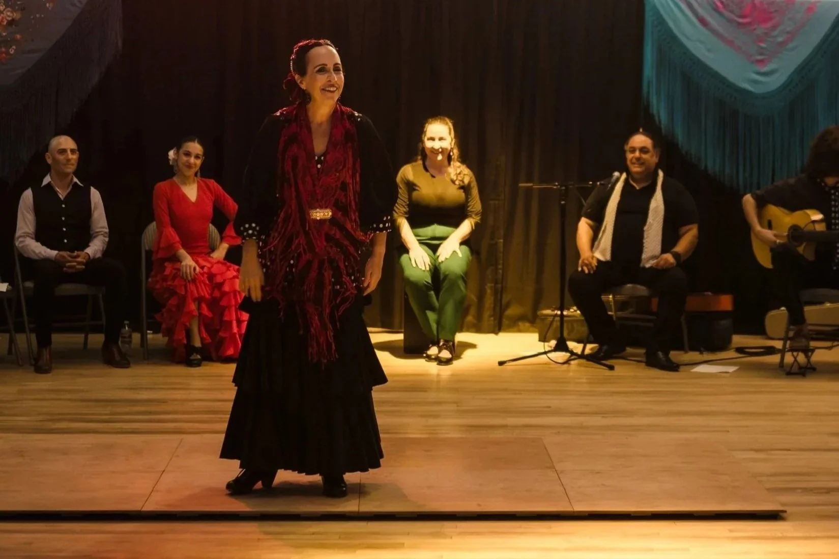 A group of performers on stage with a woman in a red dress sitting and five others seated or standing behind her, one with a guitar.