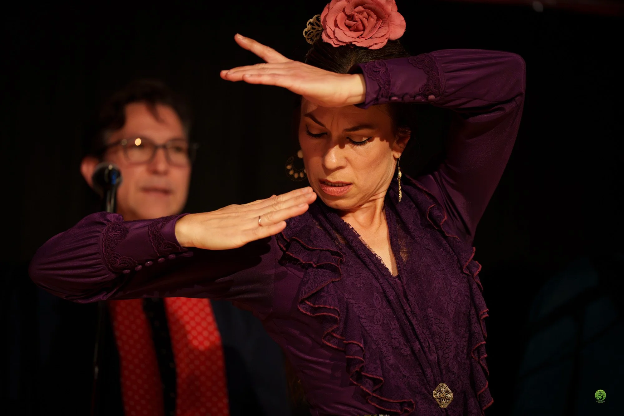A woman with dark hair wearing a purple dress with lace details is performing a dance with her eyes closed and hand above her head. A man with glasses and a red tie is blurred in the background.