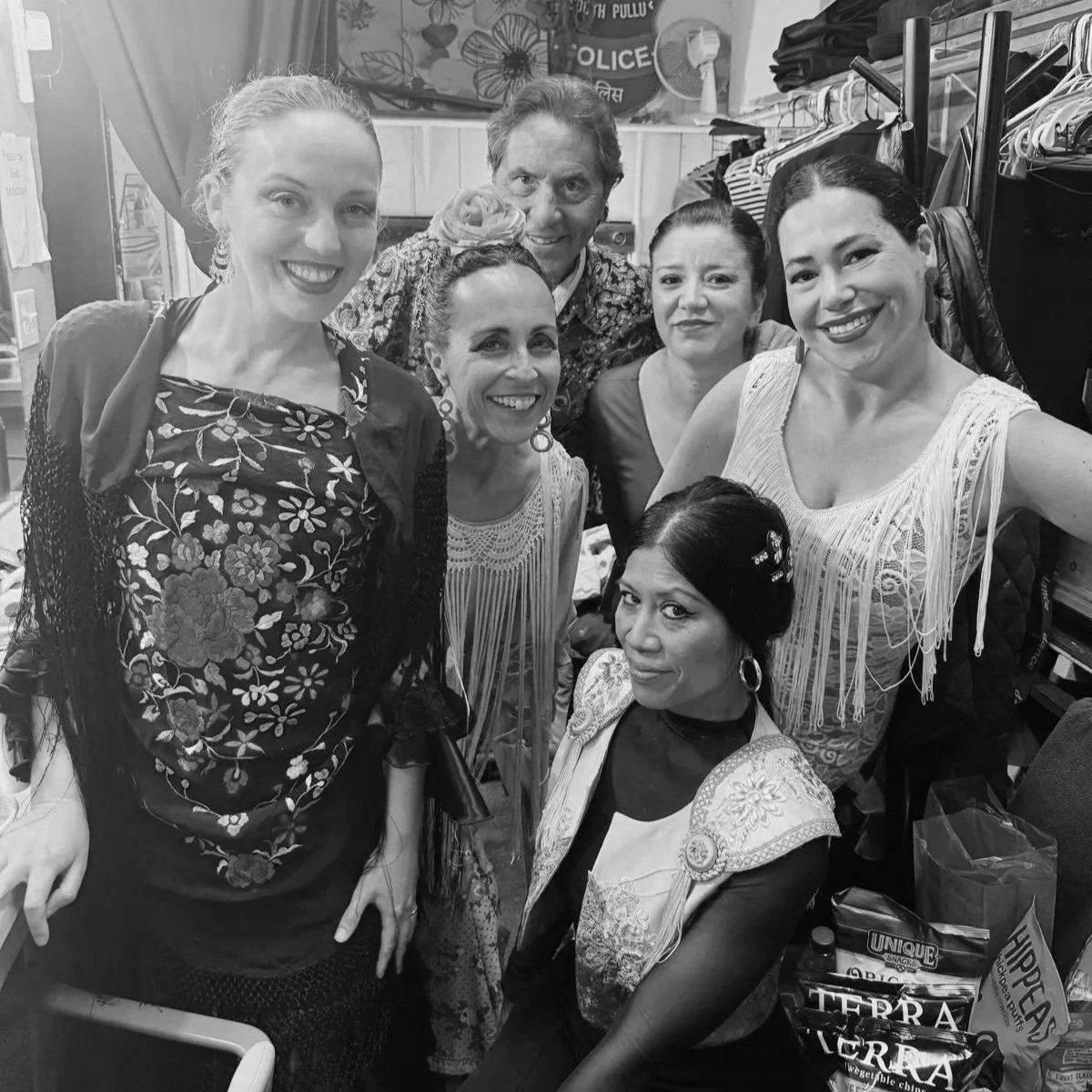 Group of five women and one man smiling indoors, in a dressing room, with shelves, clothing, and boxes in the background.