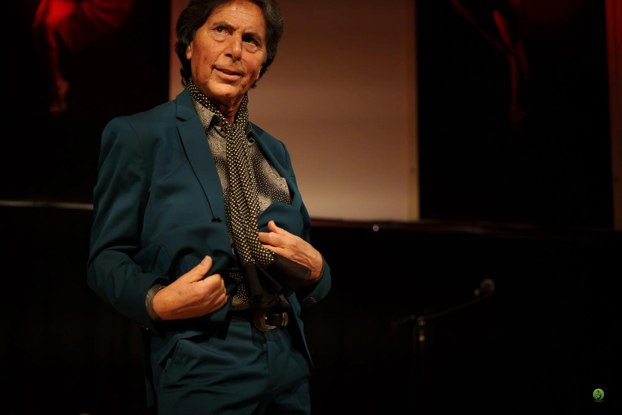 A man in a teal suit and patterned shirt sings while holding his coat.