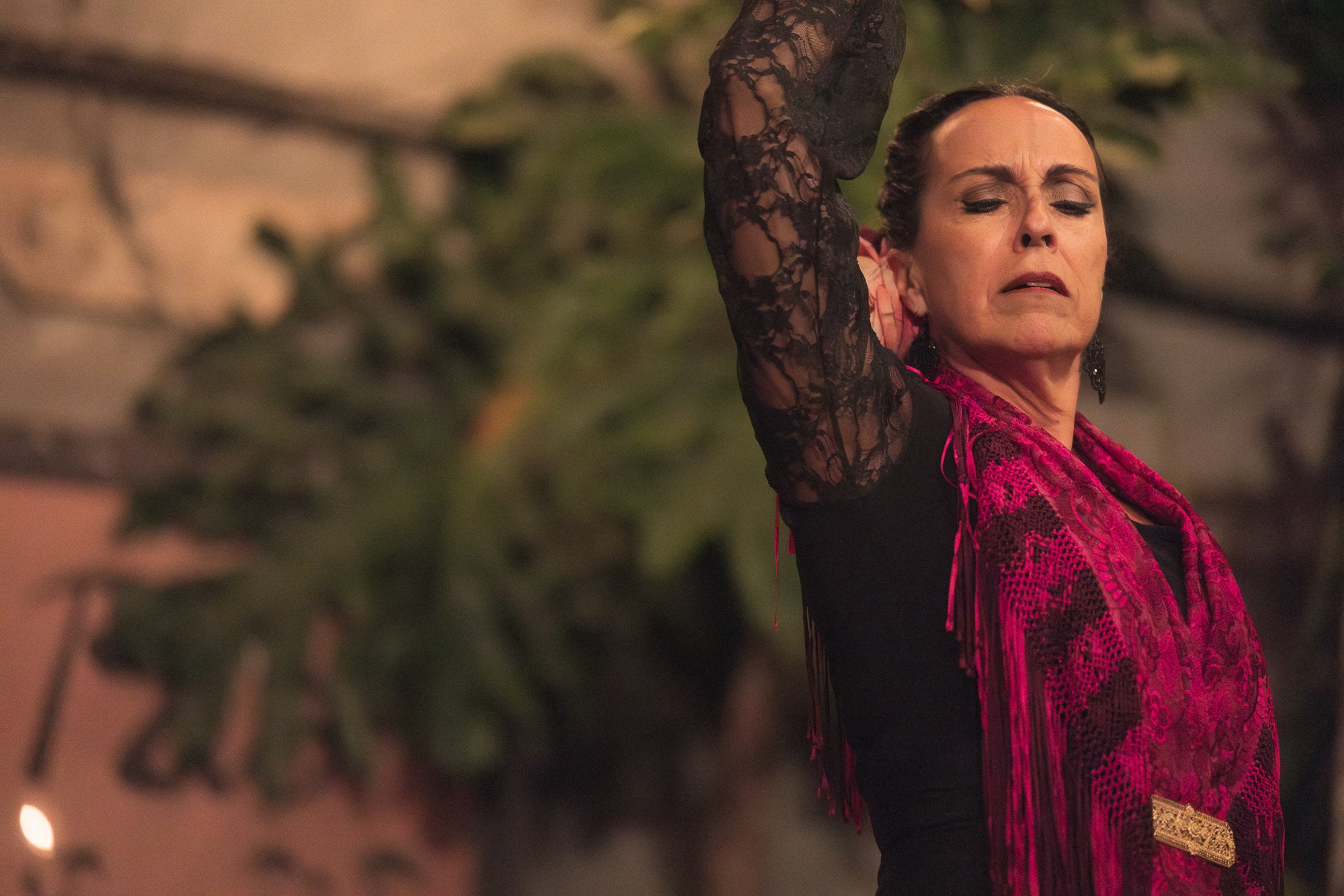 A woman with dark hair styled in an updo, wearing a black lace garment and pink shawl. She is dancing near a brick wall blurred in the foreground with some greenery and warm lighting in the background.