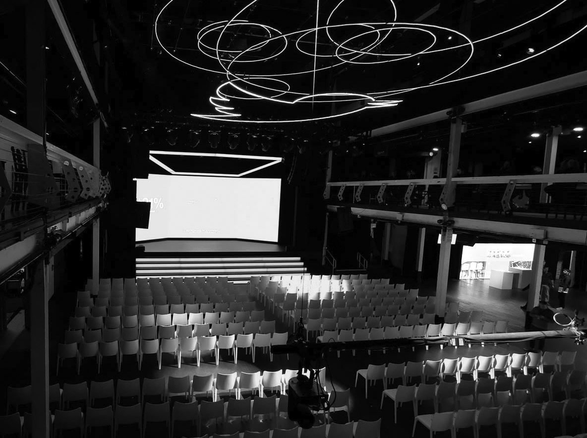 Empty conference hall with rows of chairs facing a large screen, modern ceiling lights, and two levels of balcony seating.