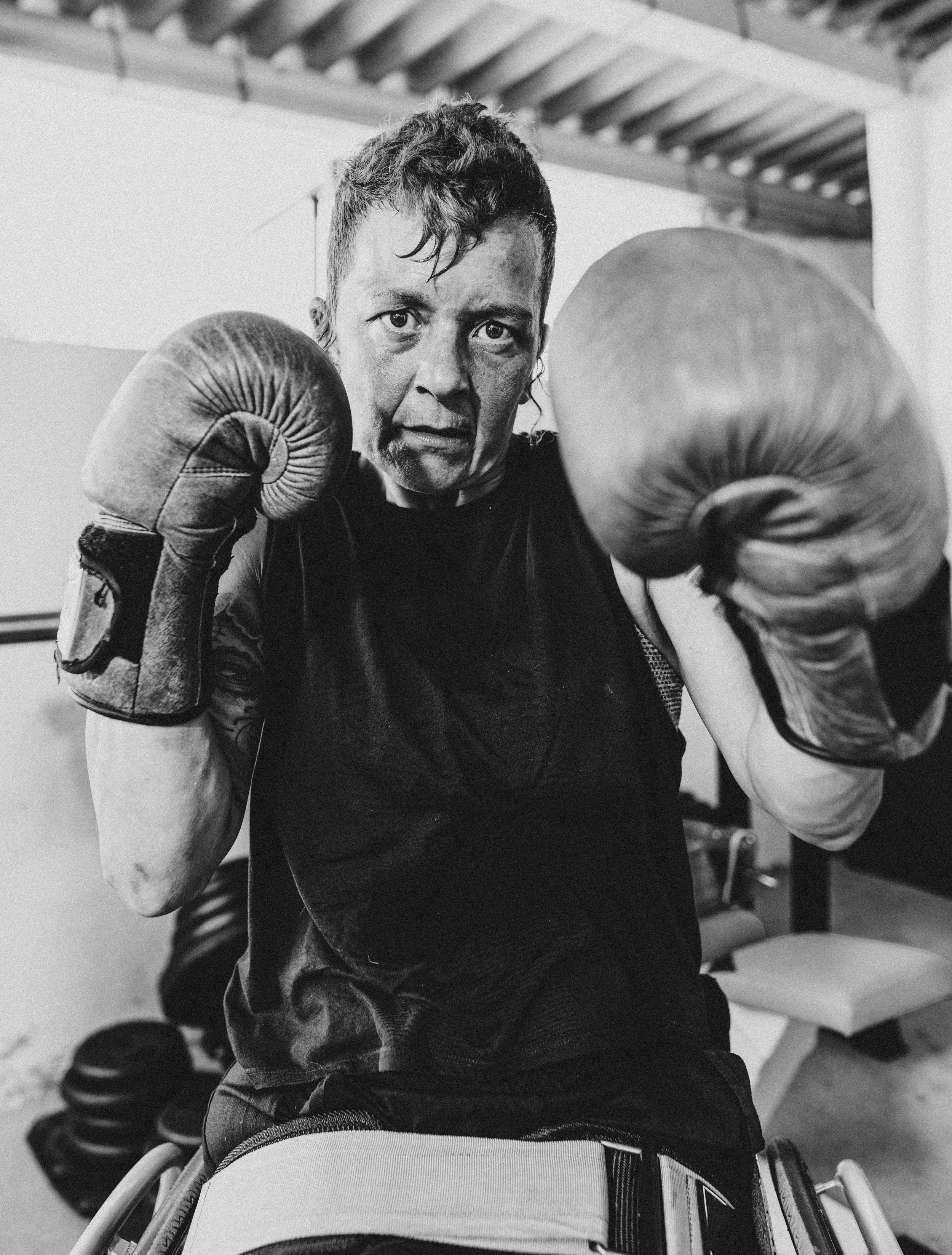 A men with short, curly hair wearing boxing gloves looks into the camera, standing in a gym.