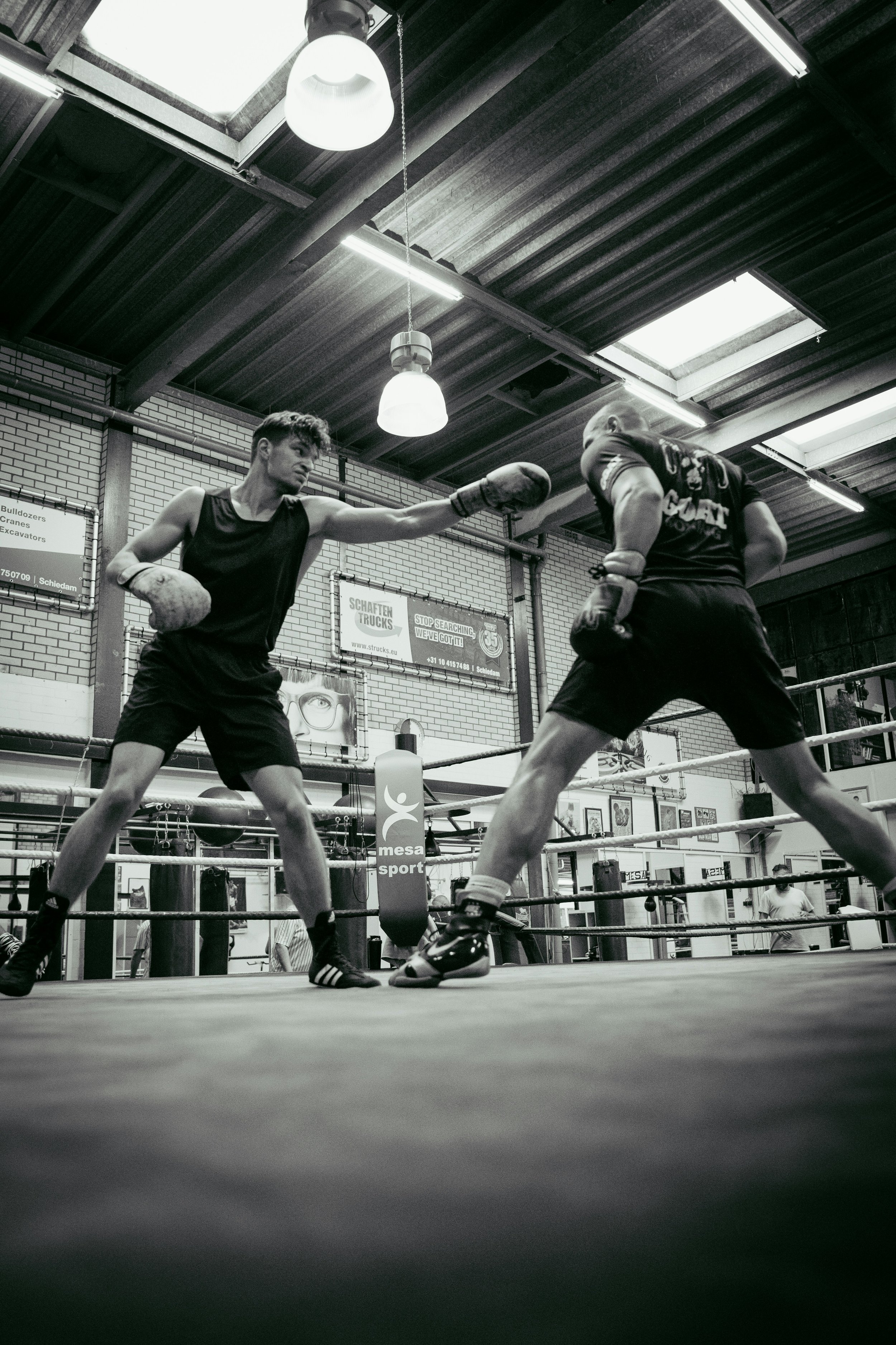 Two young men are boxing in a gym, with one throwing a punch at the other. The gym has a brick wall, ceiling lights, and posters in the background.