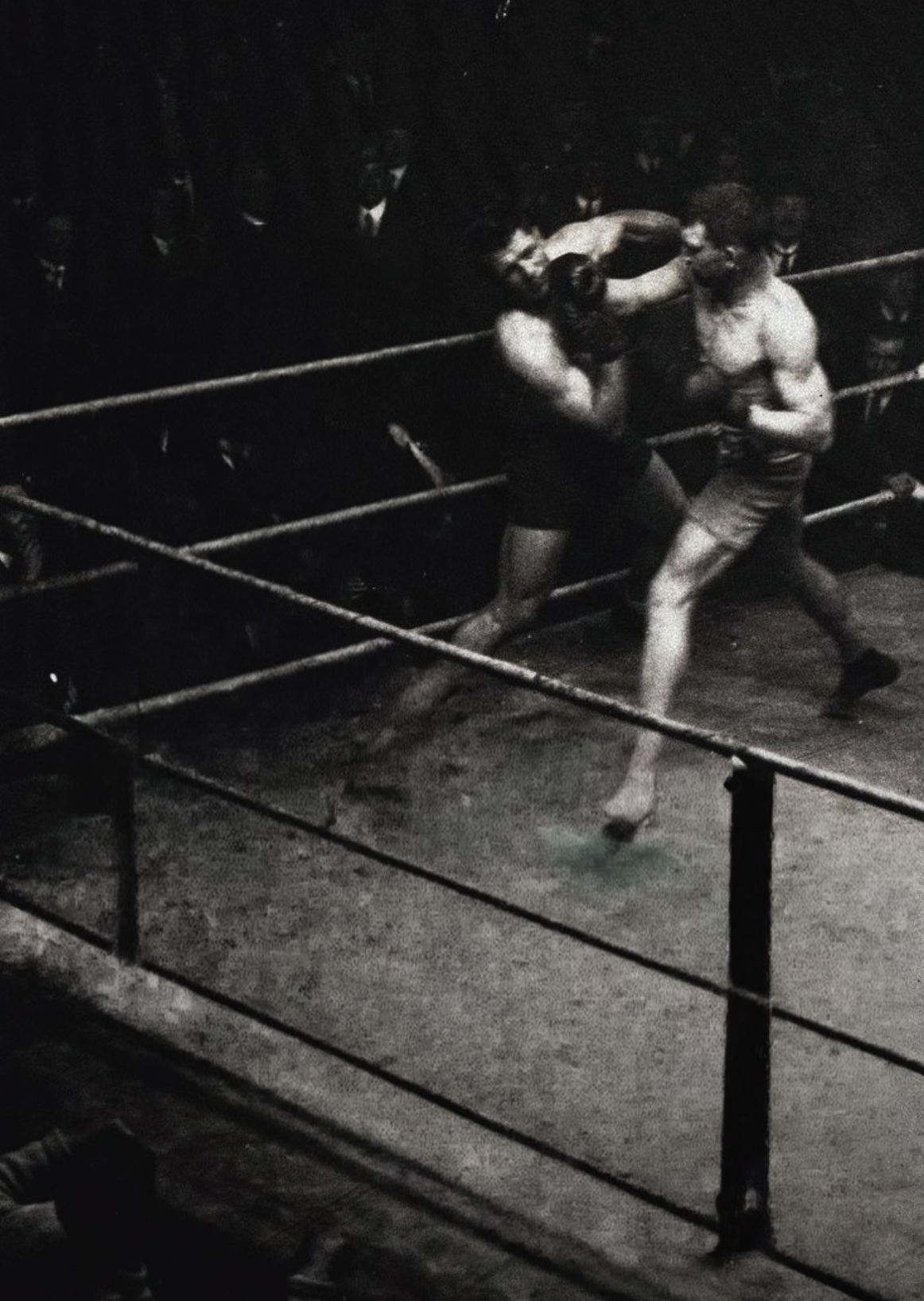 Black and white photo of two men boxing in a ring with an audience watching in the background.