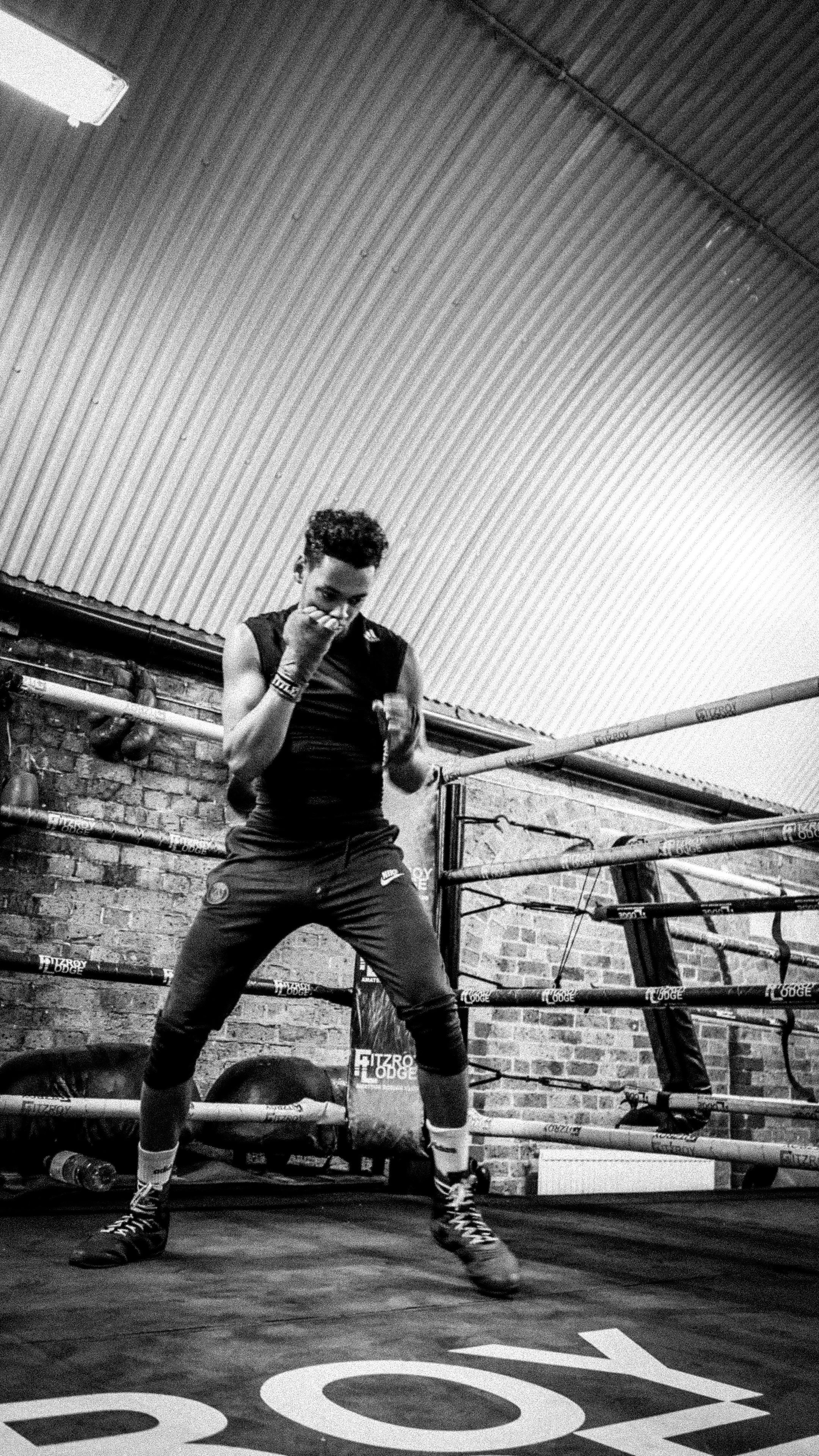 A woman boxer inside a boxing ring, wearing athletic clothing, gloves, and boxing shoes, practicing punches. The boxing ring is surrounded by ropes, and the background features a brick wall with a corrugated metal ceiling.