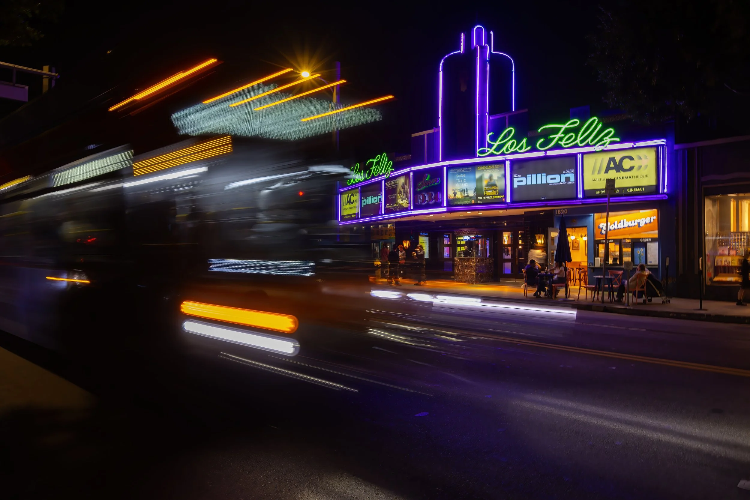 Night scene with neon signs and blurred moving lights in front of a theater called Los Fells, with outdoor seating at a restaurant.