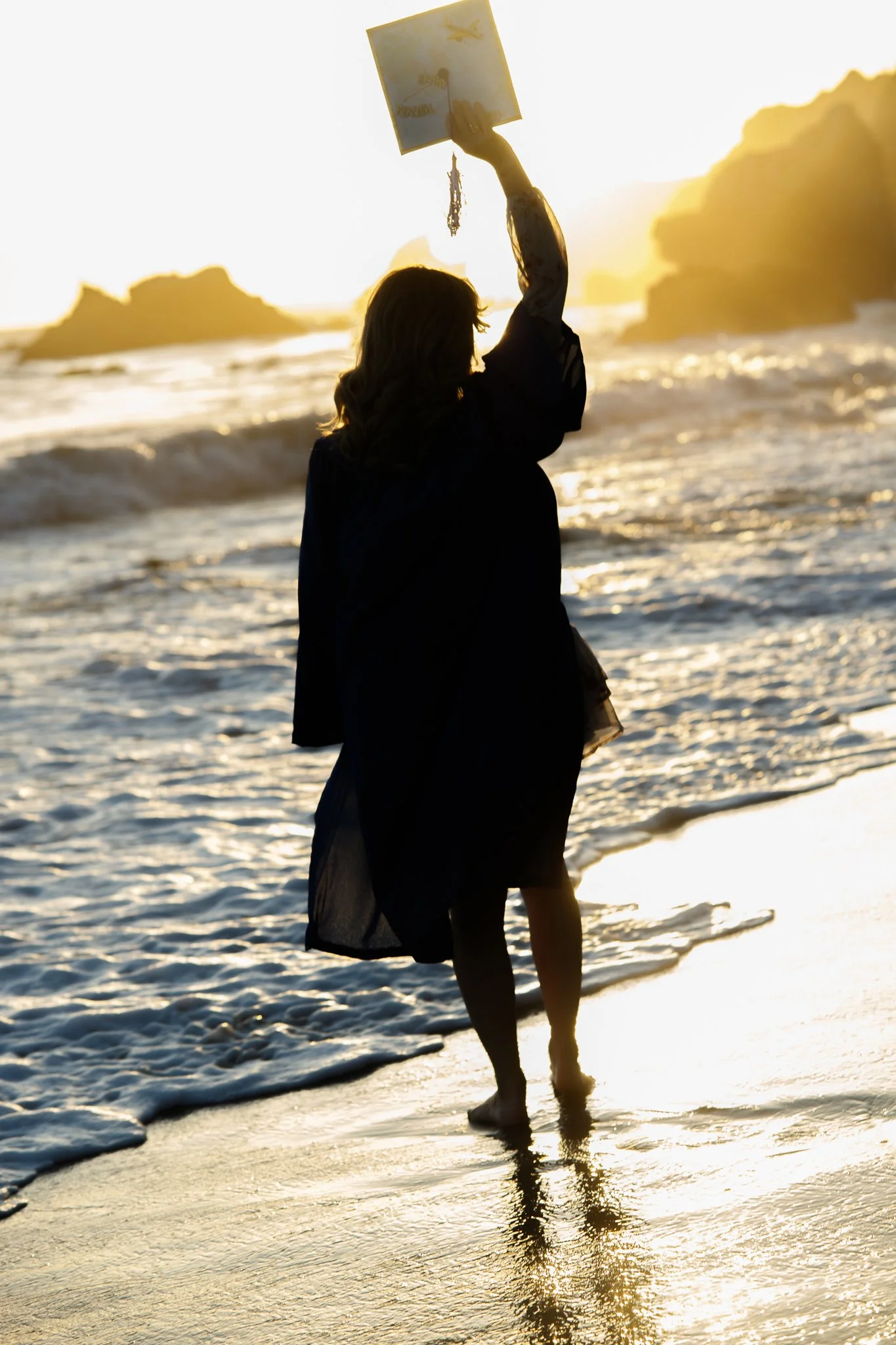 Silhouette of a woman holding a notebook with tassels at the beach during sunset, with waves and rocky formations in the background.
