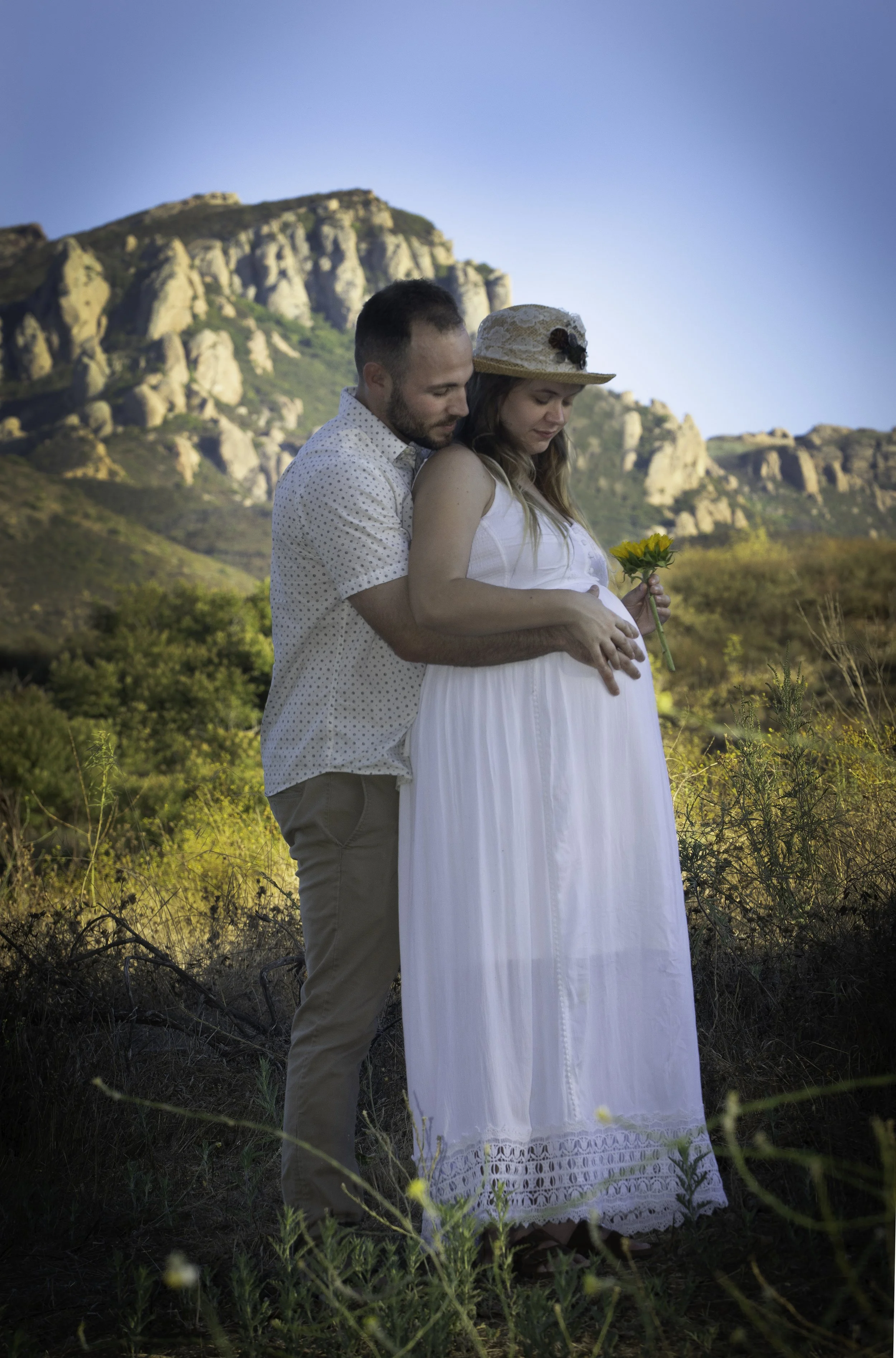 A pregnant woman in a white dress and sun hat holding a yellow flower, standing with a man in a short-sleeved button-up shirt in a natural outdoor setting with mountains in the background.