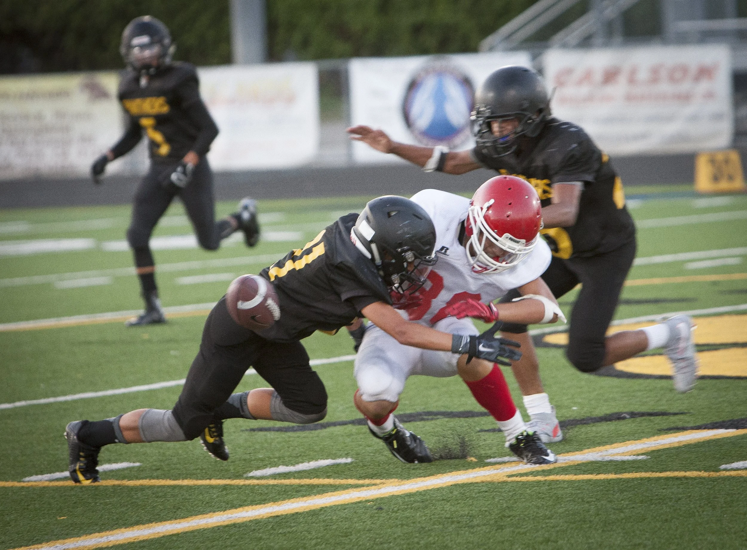 Football players in black and yellow uniforms tackle a player in a white and red uniform during a game on a field with yellow and white lines.