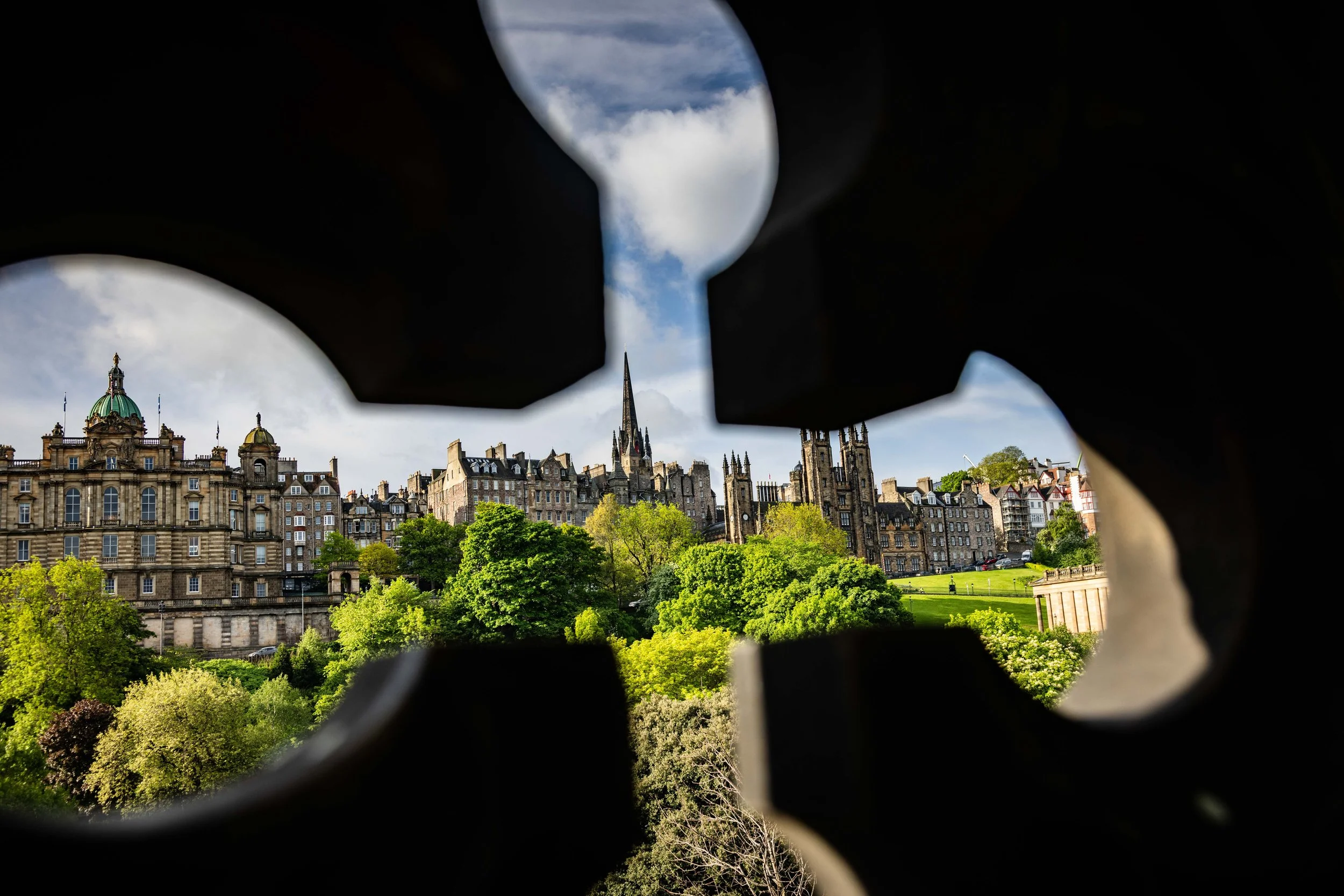 Cityscape viewed through a hole in a piece of dark material, showing historic buildings, church spires, green trees, and a cloudy sky.