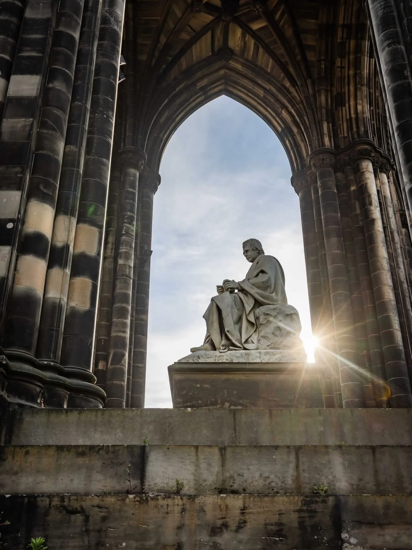 A stone statue of Abraham Lincoln seated, set against a gothic arch and dark stone columns, with the sun setting behind.
