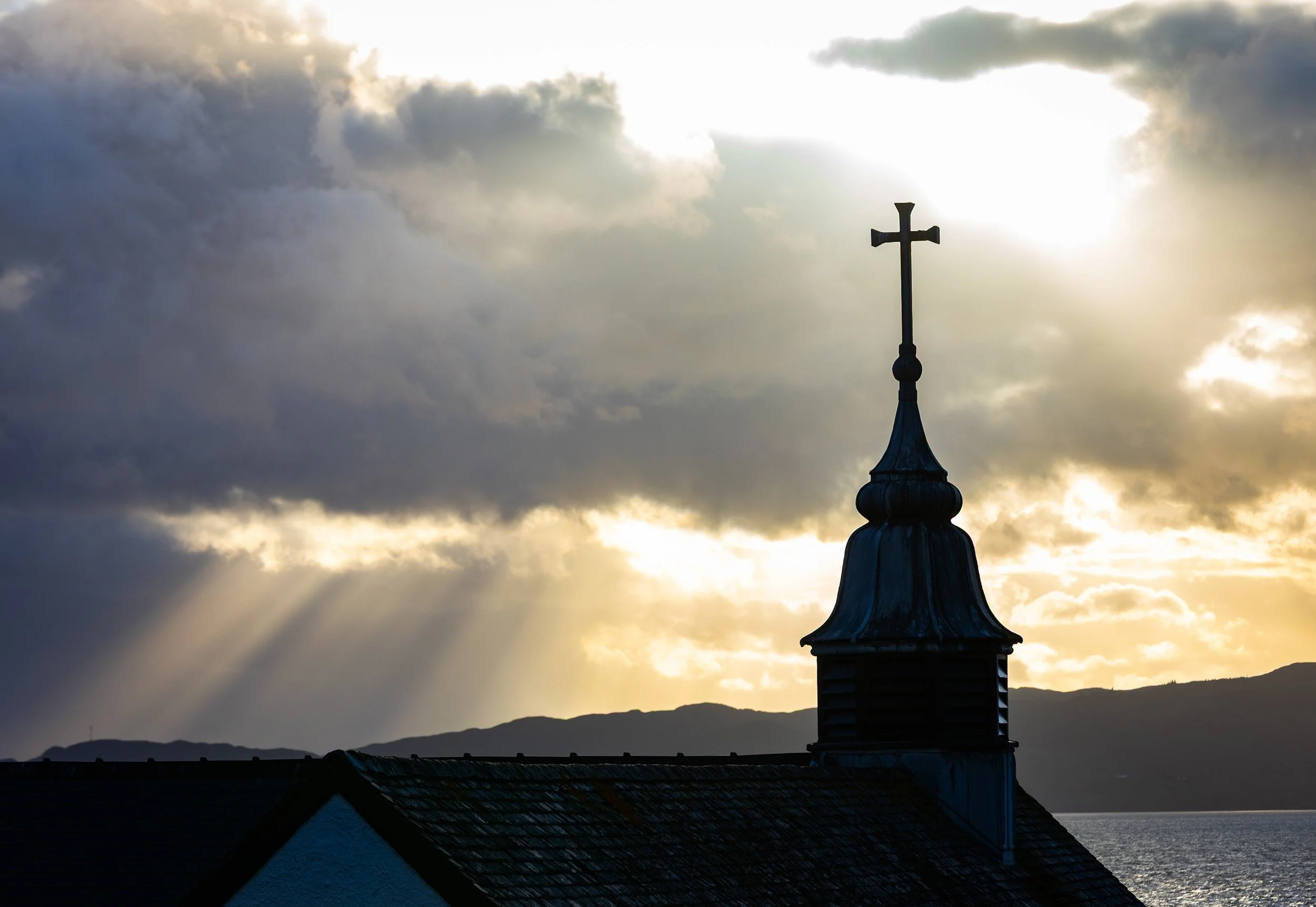 A silhouette of a church steeple with a cross on top set against a cloudy sky during sunset or sunrise.