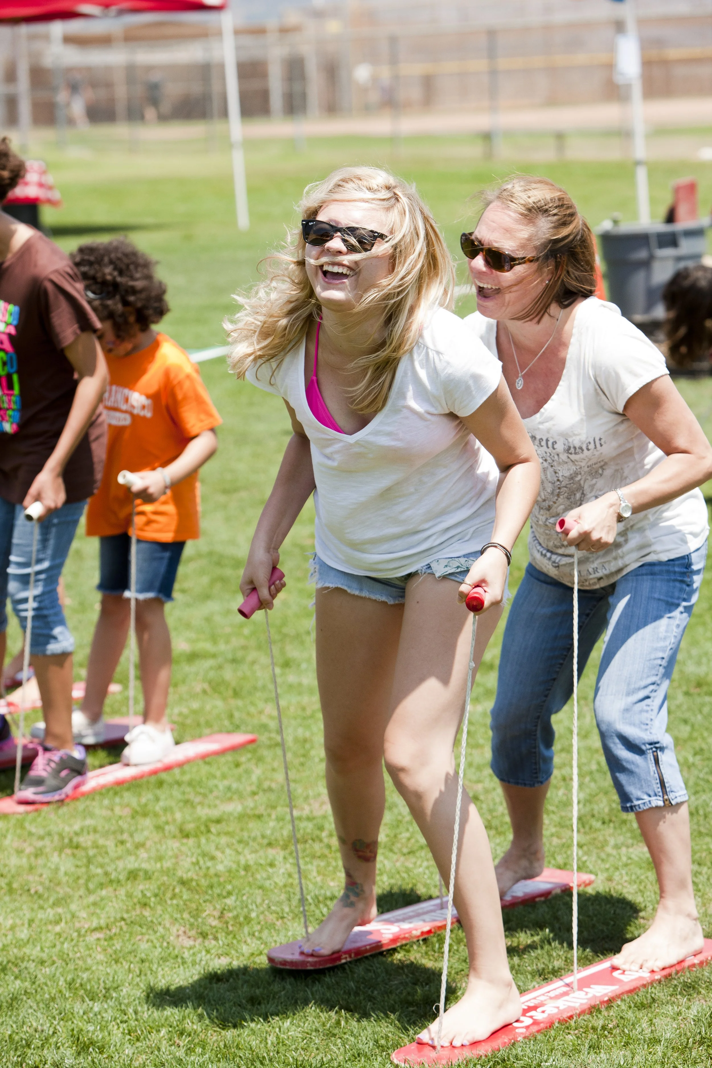 Two women and children participating in a balance activity outdoors on grass, standing on red boards with ropes, wearing casual summer clothes and sunglasses.