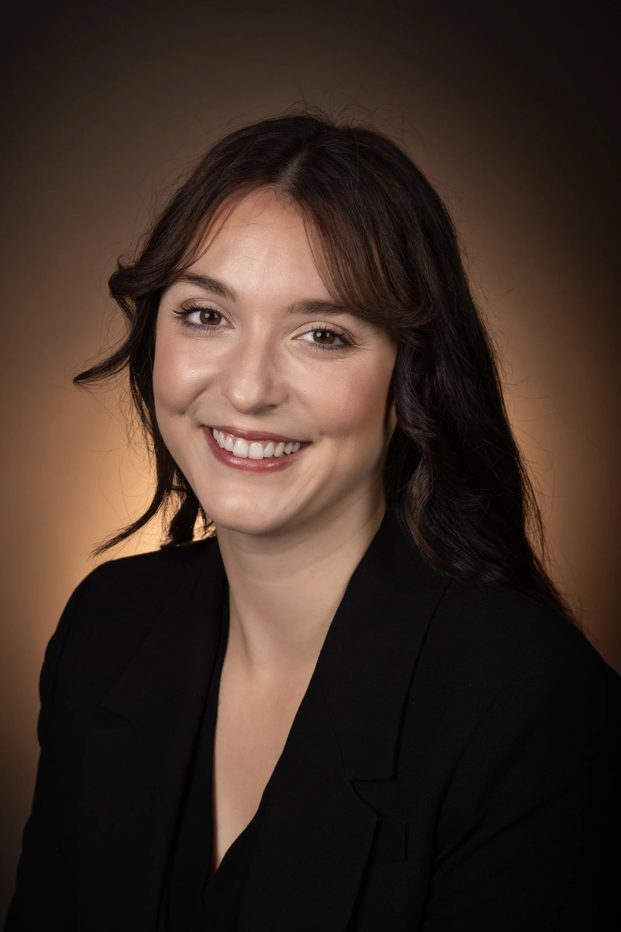 Close-up of a smiling woman with dark wavy hair, wearing a black blazer, against a brown gradient background.