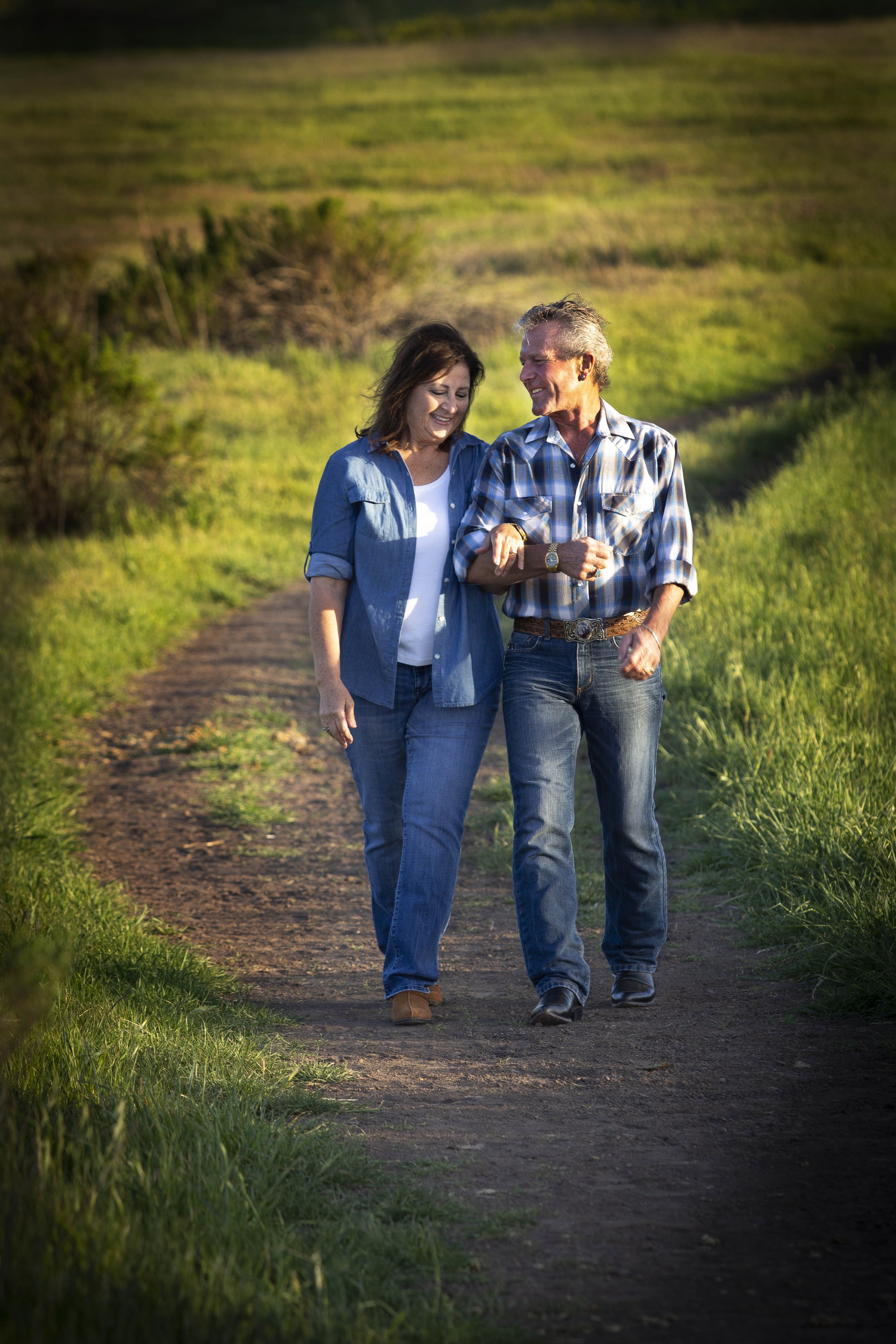 A husband and wife walking together on a dirt trail in a green field, smiling and engaging in conversation as the sun sets.