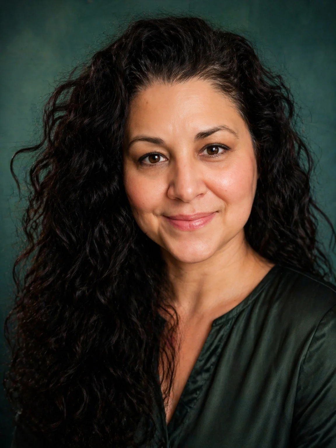 Close-up portrait of a woman with long curly dark hair, smiling, wearing a dark green top, against a dark green background.