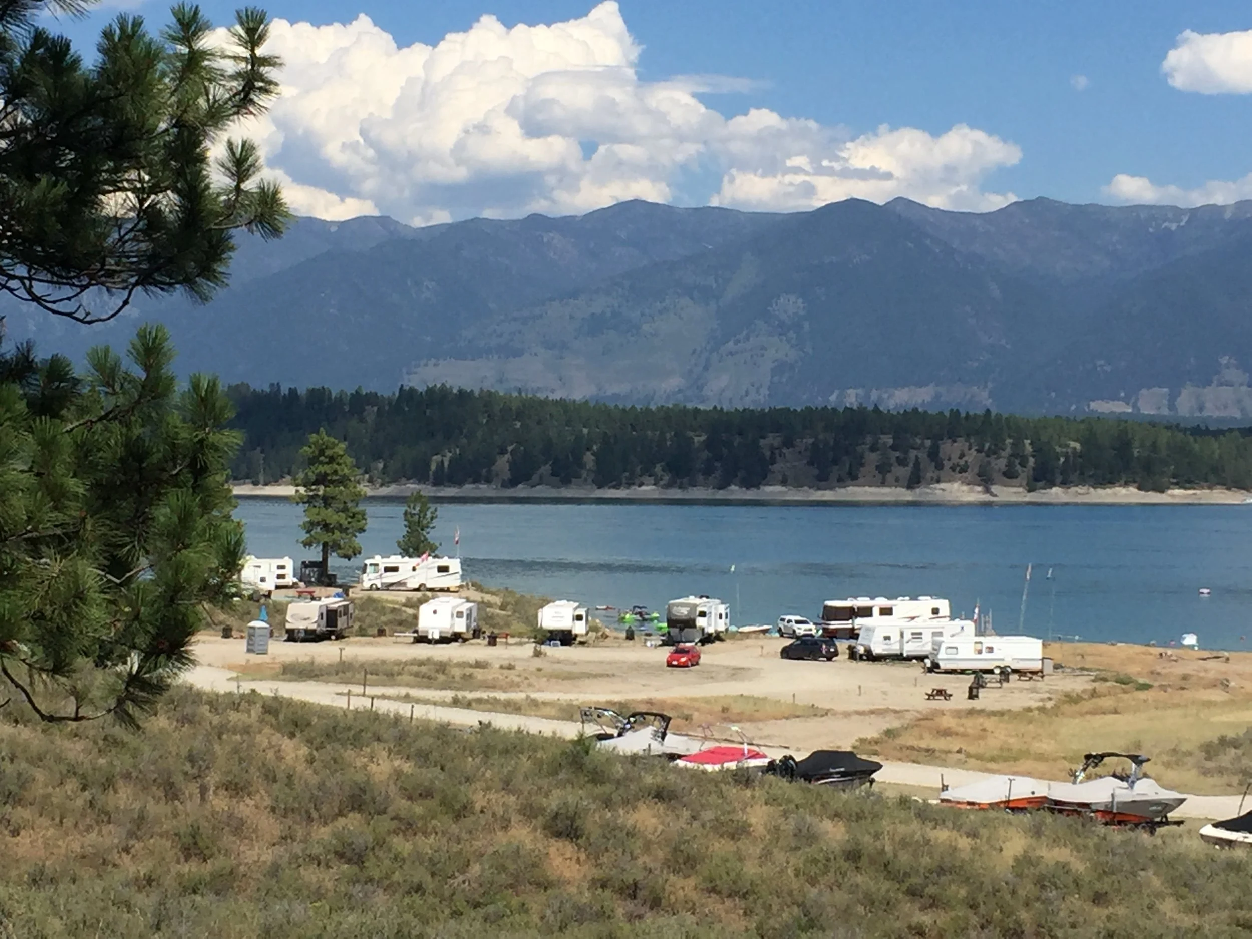 A lakeside campground with RVs, boats, and picnic tables, surrounded by trees, with mountain ranges and clouds in the background.