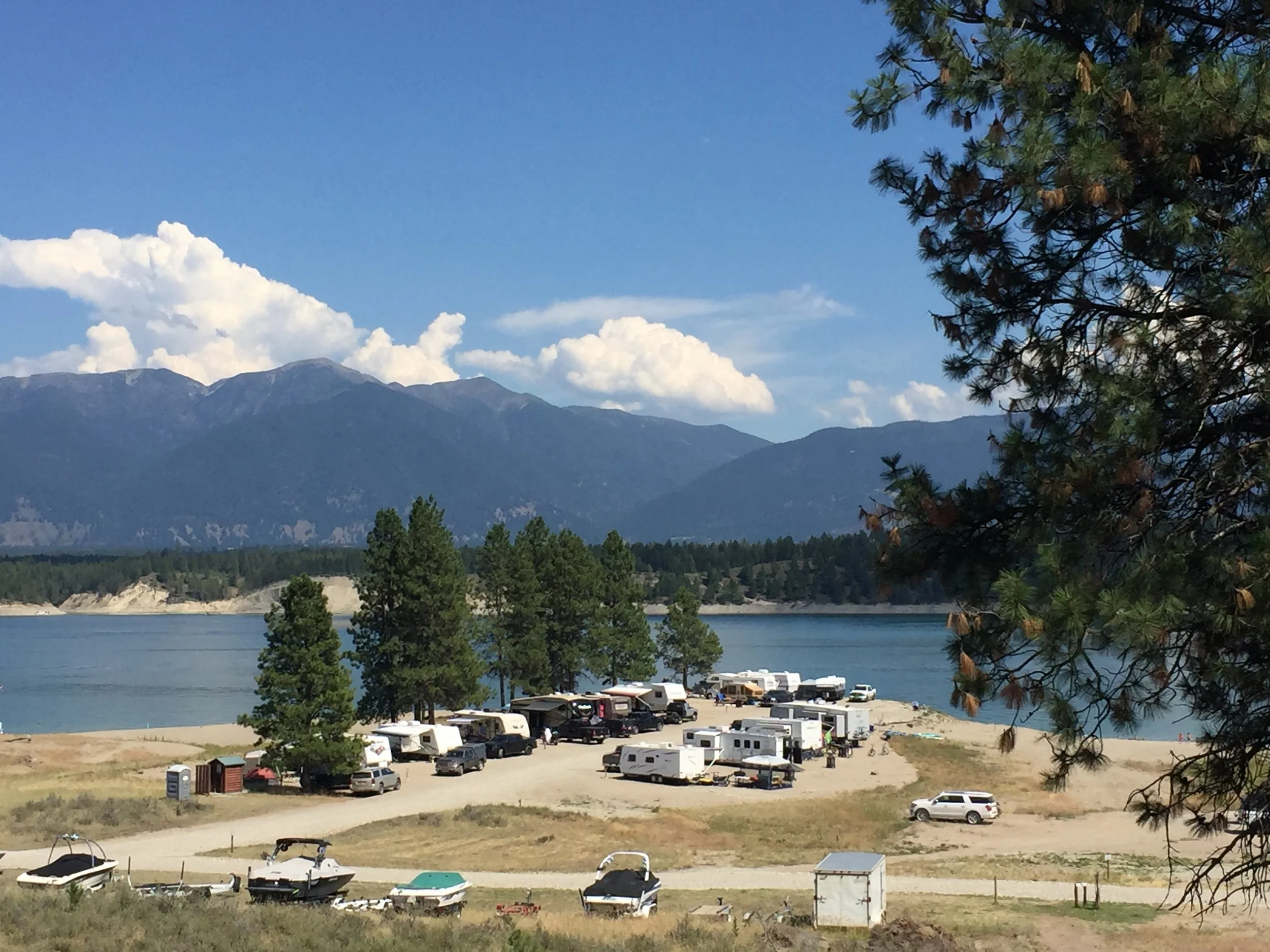 A lakeside scene with boats, RVs, and a campground set against distant mountains and a blue sky with clouds.
