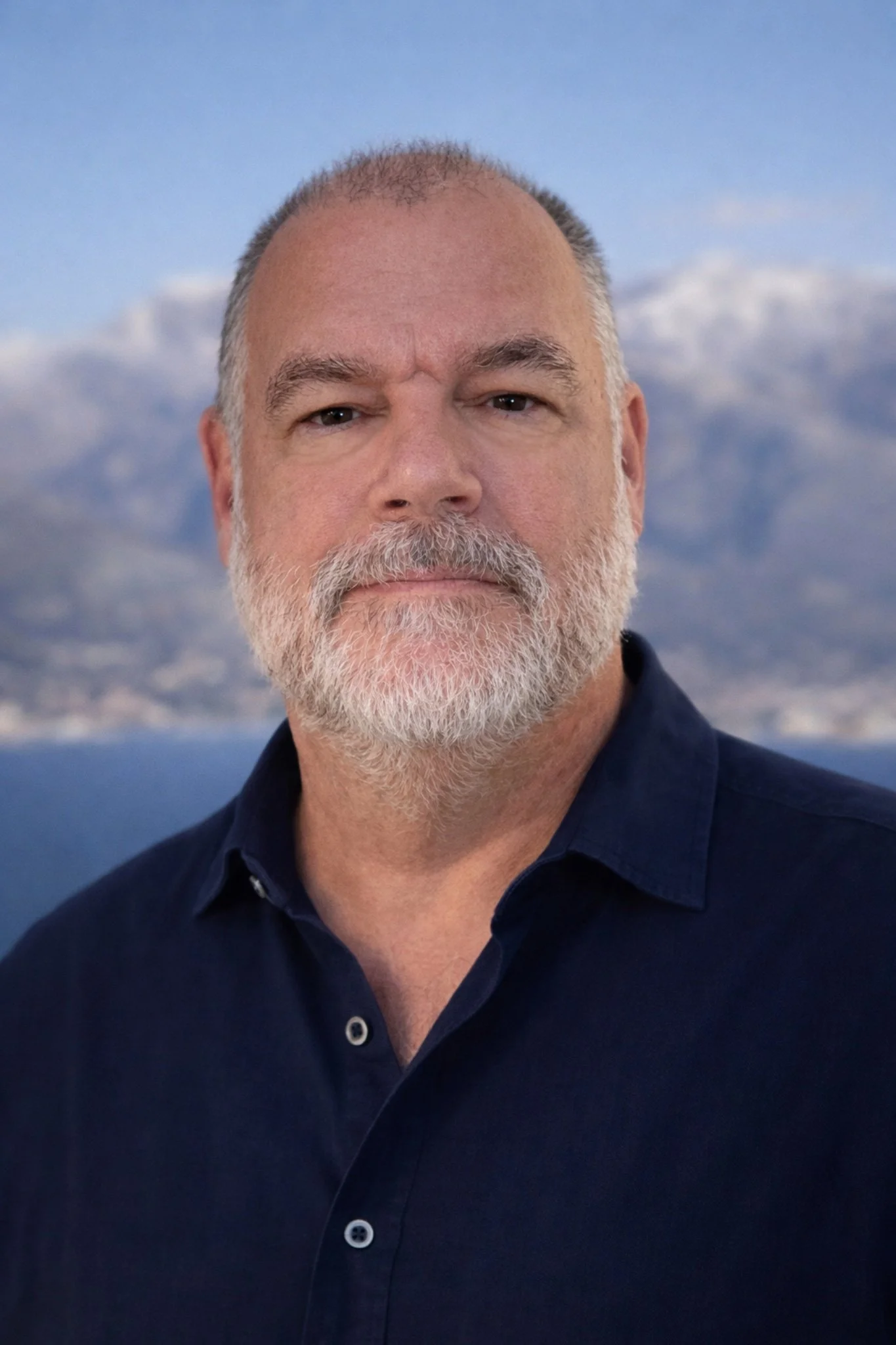 Close-up of a middle-aged man with short gray hair and a gray beard, wearing a navy blue collared shirt, against a background of snow-capped mountains and a blue sky.