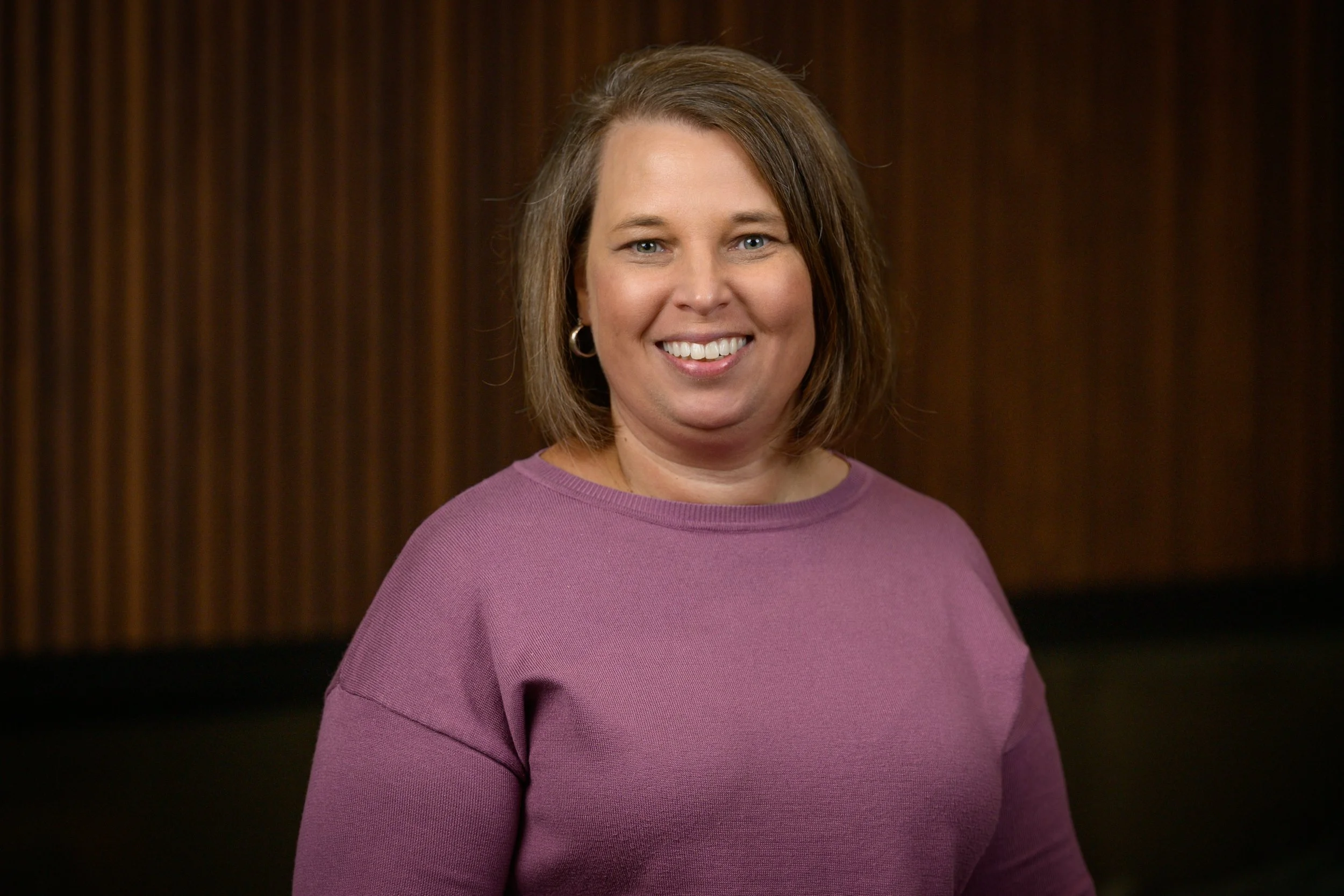 A woman with shoulder-length light brown hair and blue eyes, smiling, wearing a purple long-sleeve shirt, against a dark wooden background.