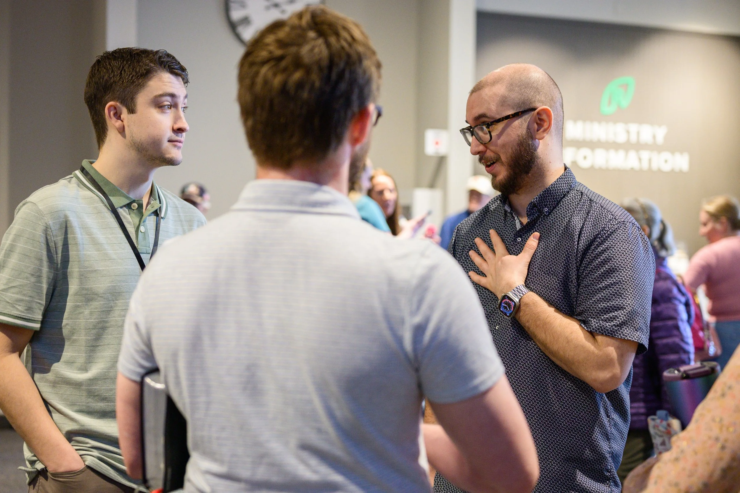 Three young men engaged in conversation at a public event, with a blurred background showing more attendees and a wall sign reading 'Ministry of Information'.