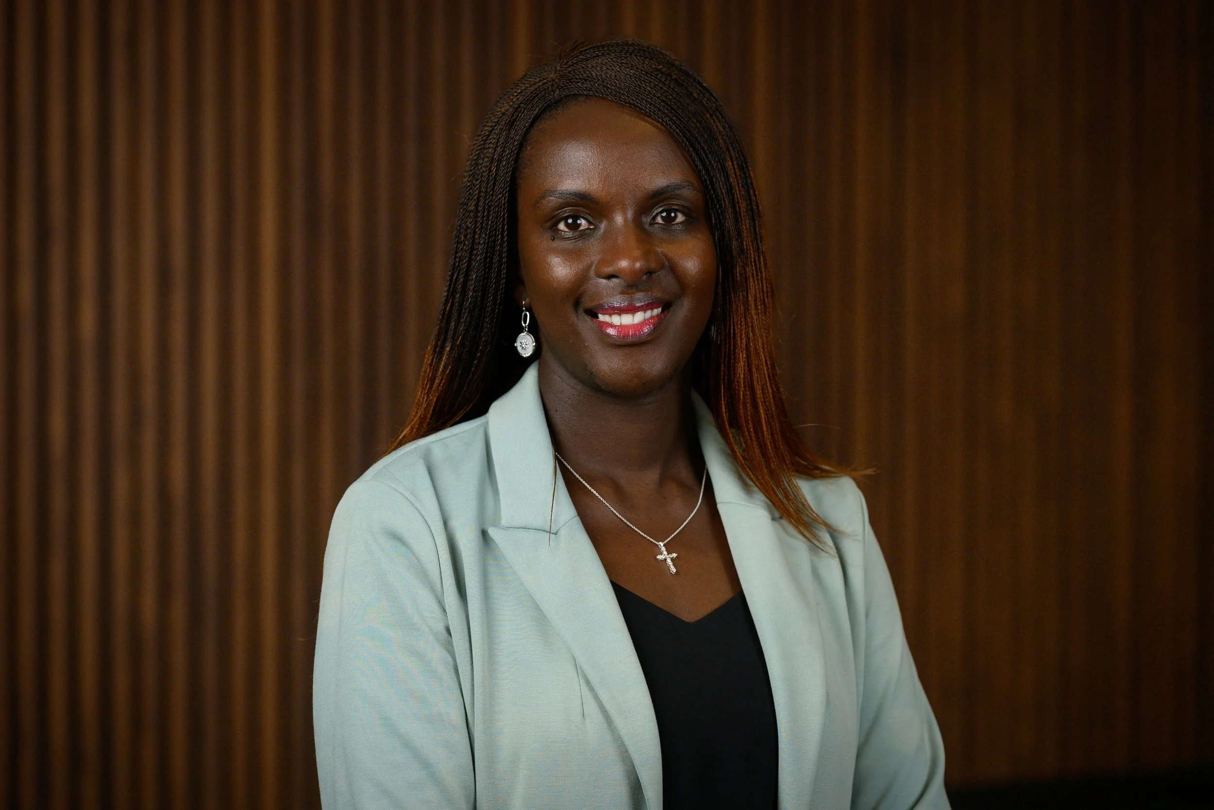 Portrait of a smiling Black woman with long braided hair, wearing earrings, a silver cross necklace, a black top, and a light green blazer, standing against a brown wood-paneled background.
