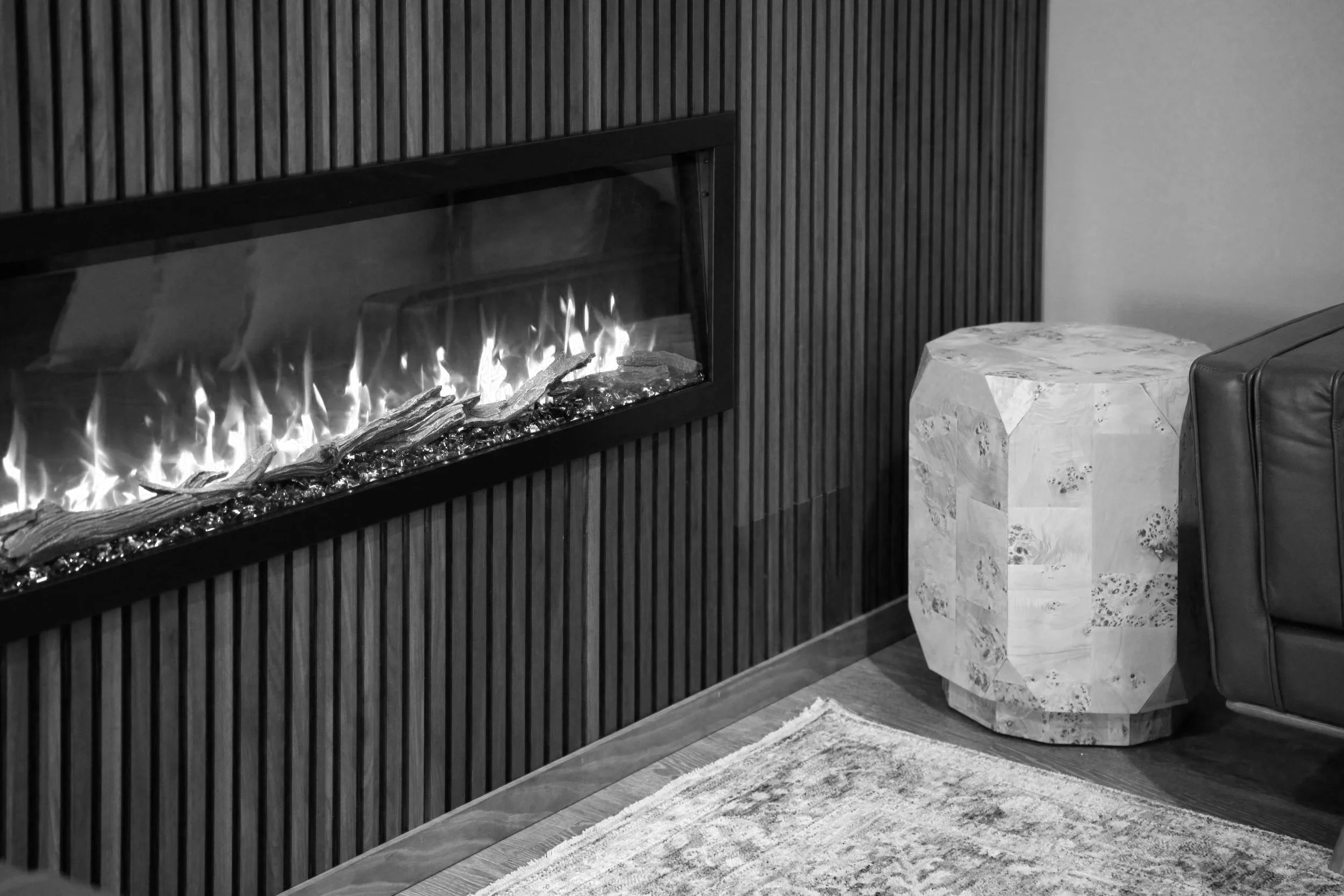 Modern living room corner with a black-framed glass fireplace built into a black vertical slatted wall, a marble side table, a dark sofa, and a light-colored rug.