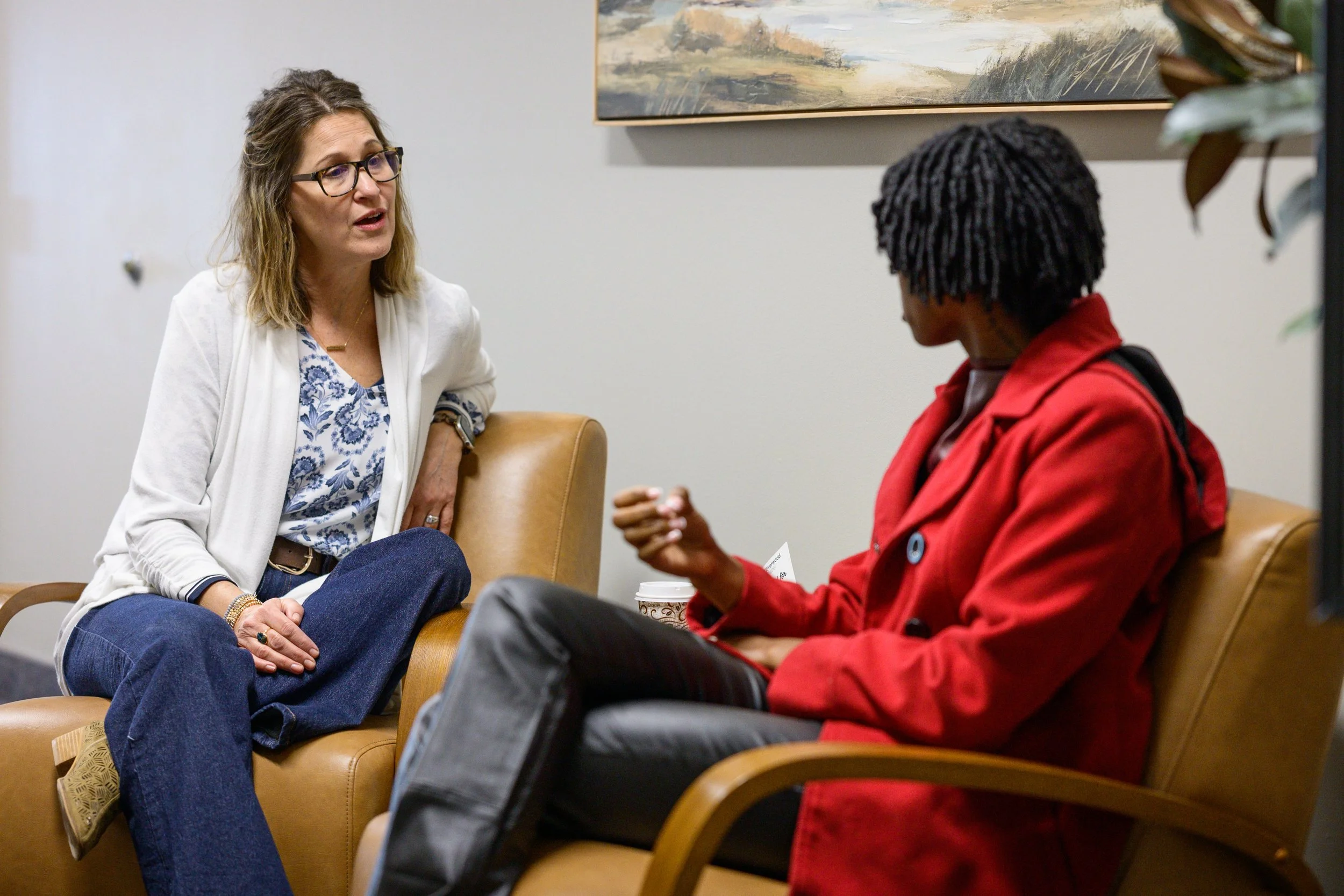 Two women having a conversation in a waiting area, sitting on tan chairs with a painting on the wall behind them. One woman has brown hair, glasses, a white blazer, and blue jeans, and the other woman has black dreadlocks, a red jacket, and dark pants.