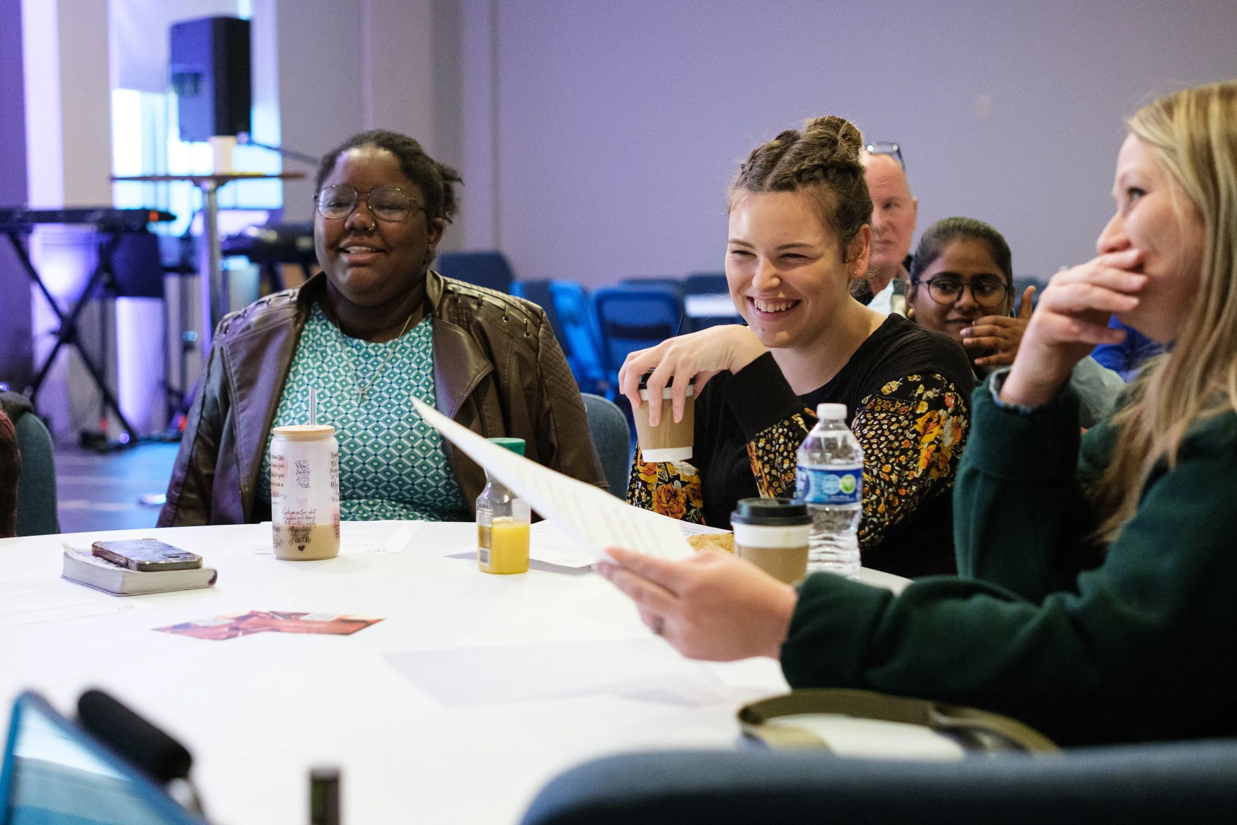 A group of people sitting around a table in a conference or workshop setting, engaging in conversation and laughing. The table has drinks, papers, and a book, with a stage and musical equipment in the background.